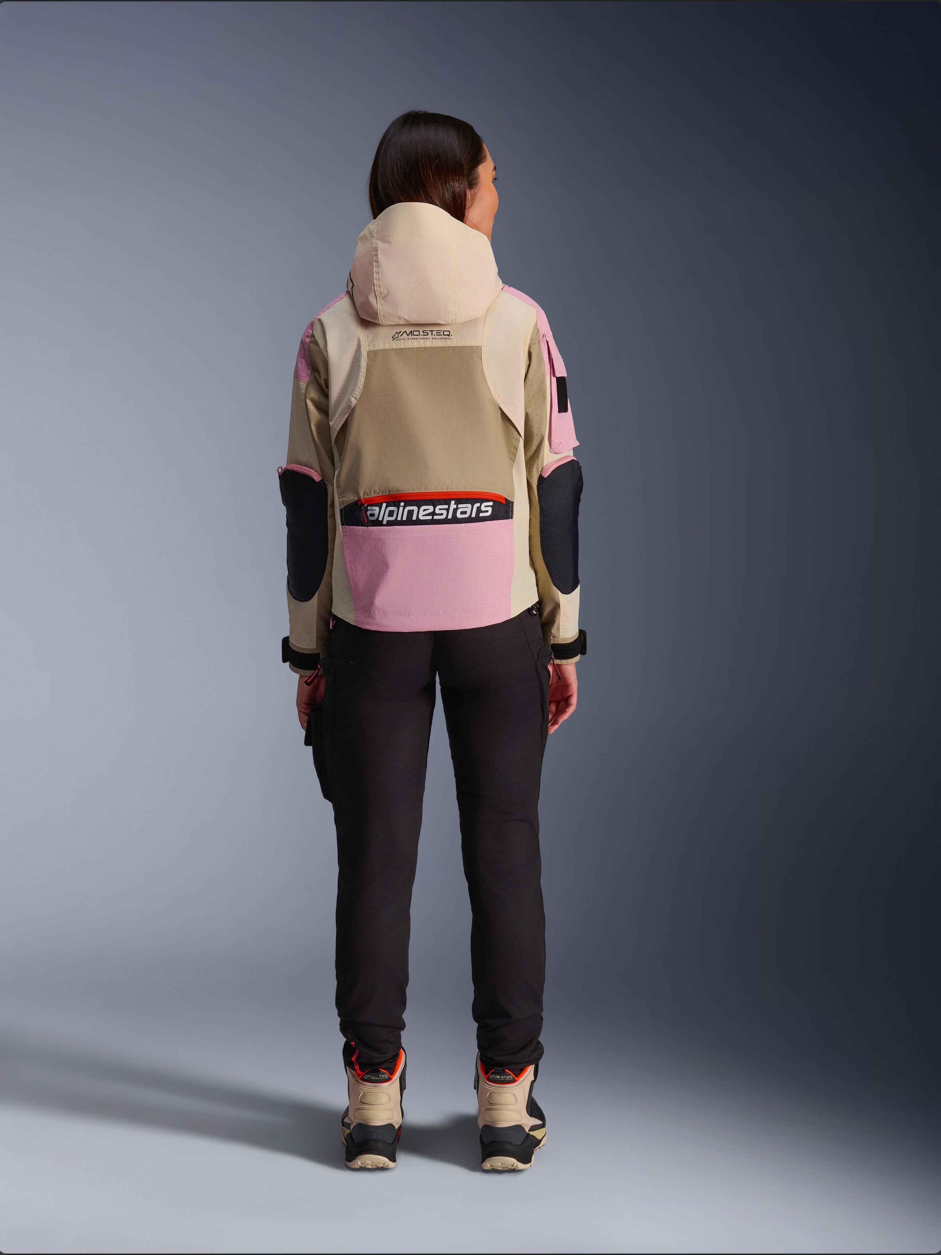 Female rider viewed from behind wearing the Alpinestars Molly Air Jacket in Sand Pink Almond, featuring a tan hood and pink panels with black accents, paired with black riding pants and tan motorcycle boots, urban contemporary motorcycle apparel
