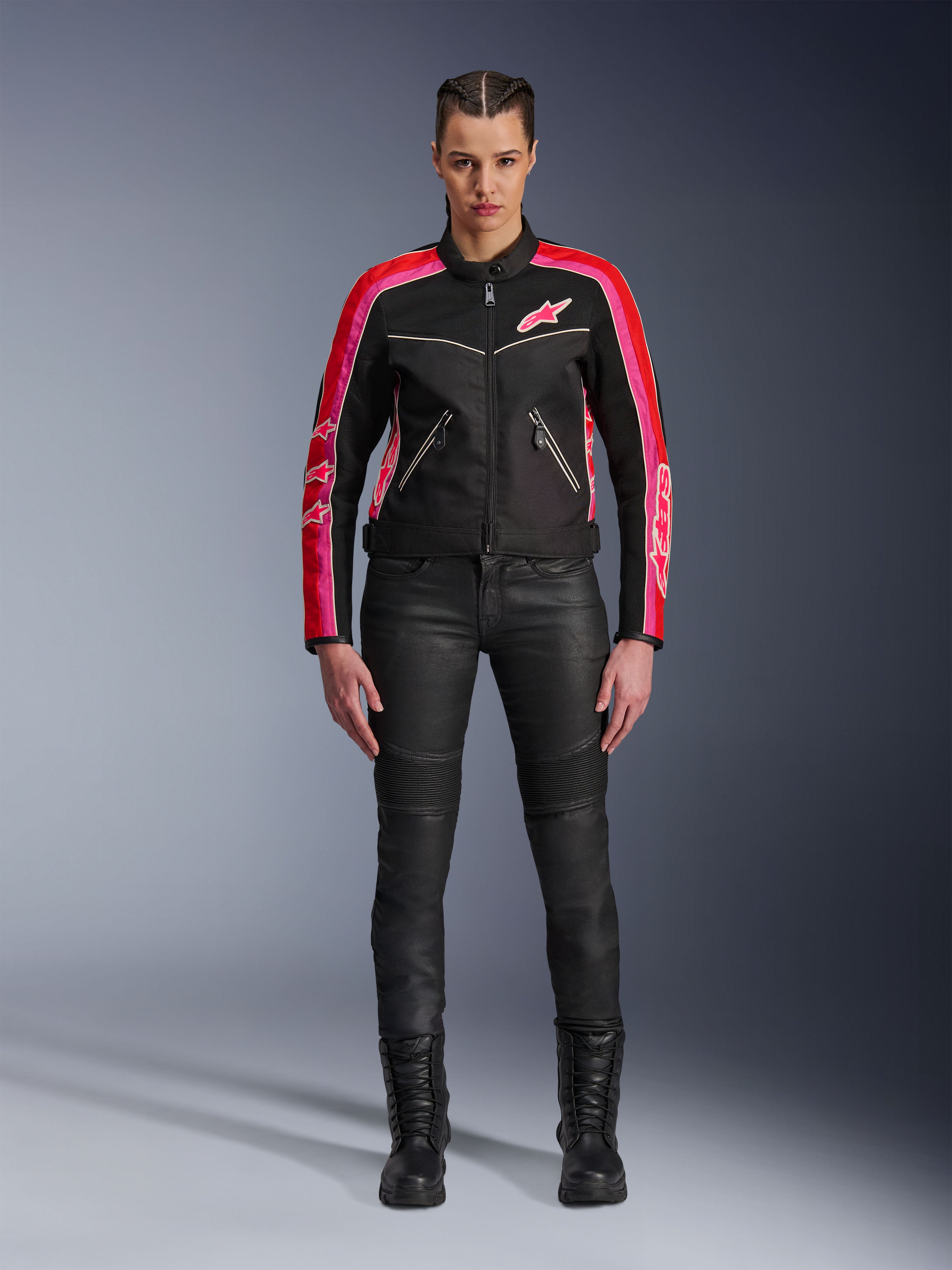 Motorradfahrerin in der Alpinestars Stella T-Dyno Air Jacke, Schwarz mit hellroten und fuchsiafarbenen Akzenten, kombiniert mit schwarzer Motorradhose und Schnürstiefeln, mit 3D-Mesh-Einsätzen, taillierter Passform und pinkfarbenen Stern-Logos, urbanes Motorrad-Outfit