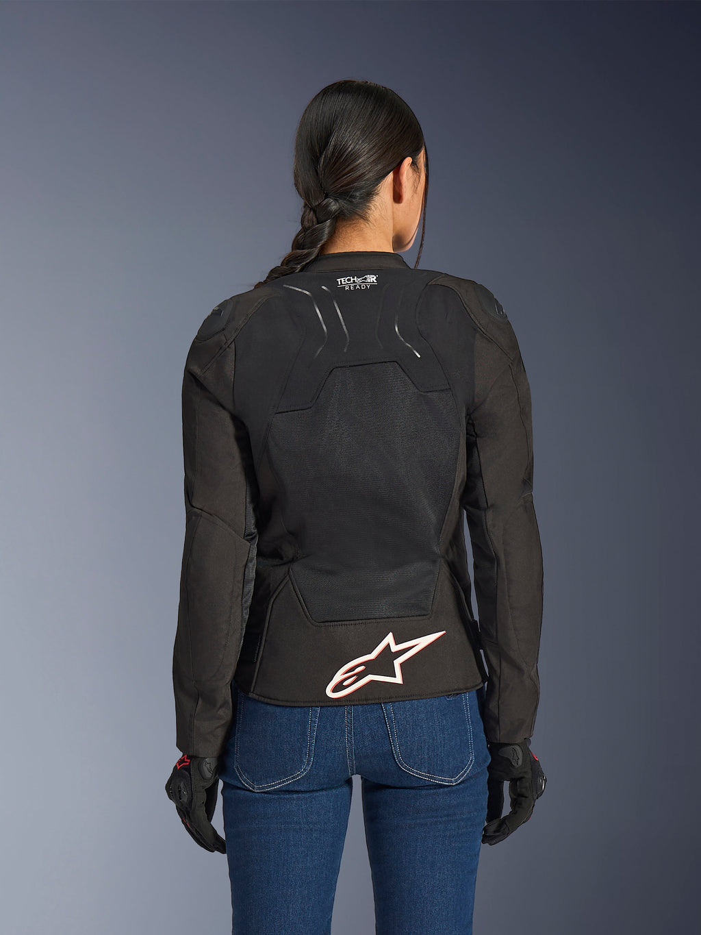 Alpinestars Stella SMX Air Jacket, back view on female model, black textile with high-flow mesh panels, 'TECH-AIR READY' branding on the upper back, large white Alpinestars logo with red outline on the lower back, paired with blue denim jeans and black riding gloves