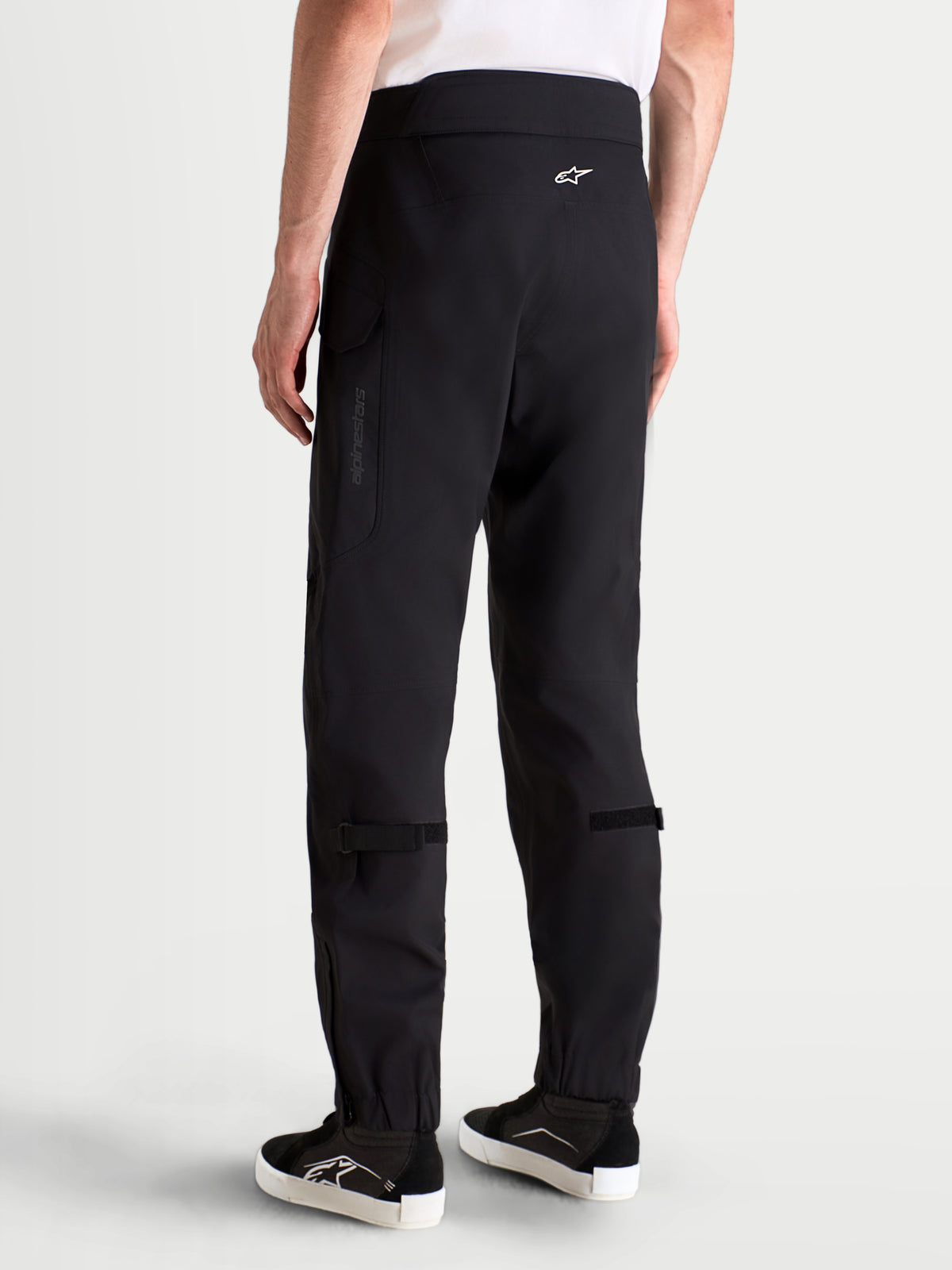 Alpinestars Juggernaut V 2 Drystar Hose, urbane wasserdichte Herren-Motorradhose in Schwarz, Rückansicht an einem Model mit passender Ausrüstung und Schuhen, mit hohem Bund und Cargotaschen