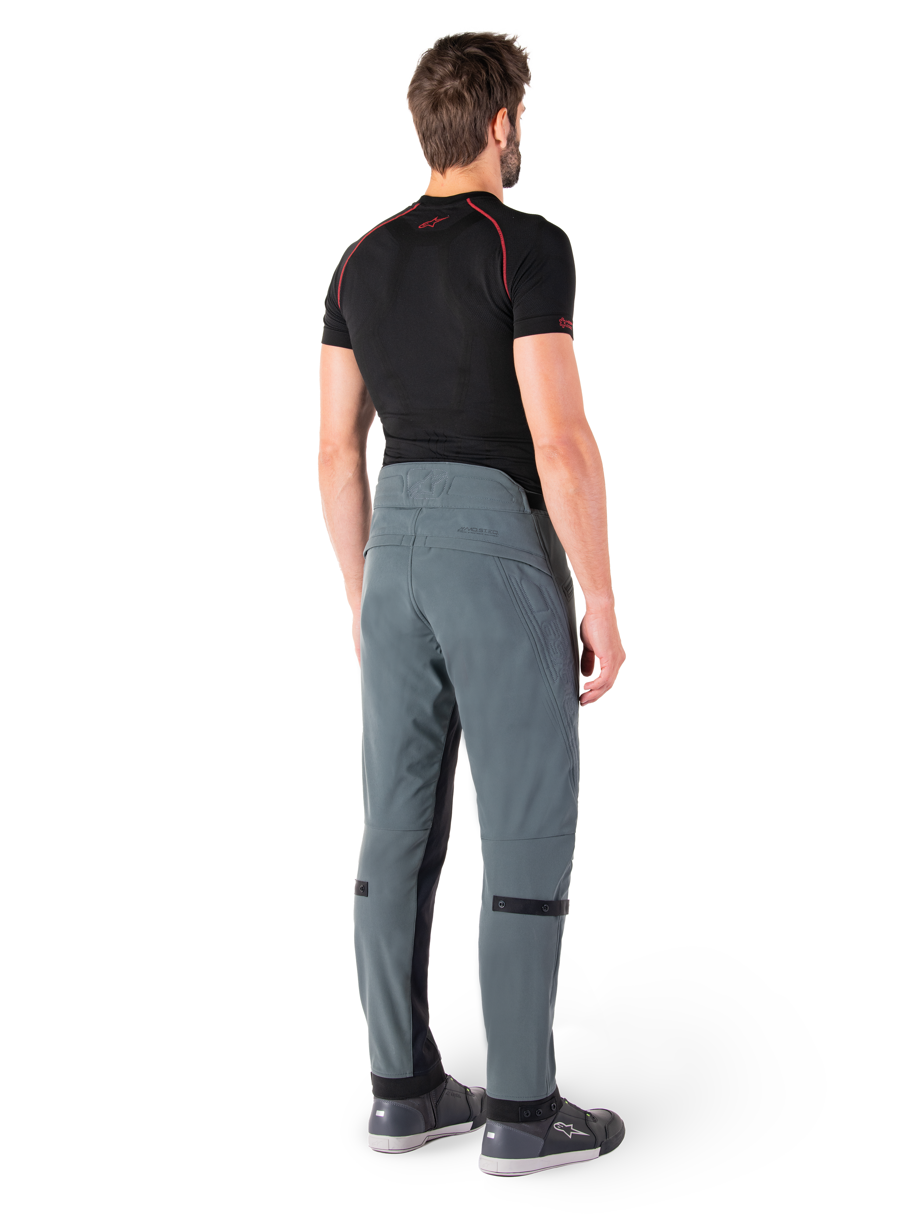 Alpinestars Alden Hose, Urban Grau, Rückansicht, technische Textil-Motorradhose mit gesteppten Details am oberen hinteren Bund und an den Beinen, kombiniert mit einem schwarzen Baselayer und grauen Motorradschuhen, moderner urbaner Streetwear-Stil.