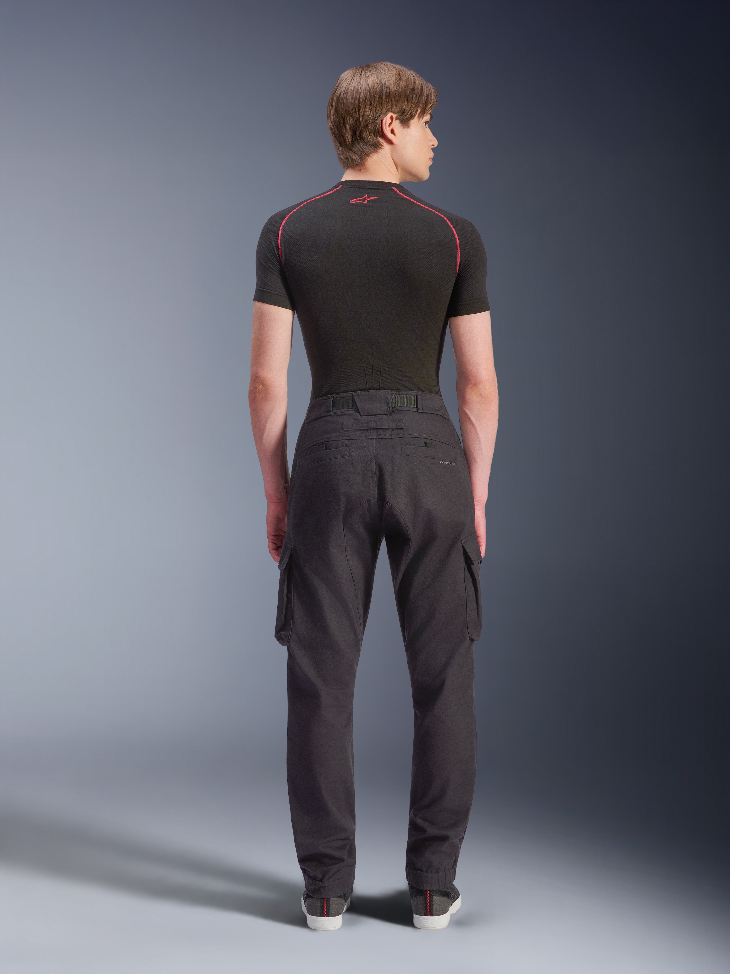 Alpinestars Flex-AST Cargo Hose, Motorrad-Cargohose, Schwarz, Rückansicht am Modell, mit regulärer konischer Passform, elastischen Säumen und seitlichen Cargotaschen, kombiniert mit einem schwarzen Baselayer und lässigen Sneakern