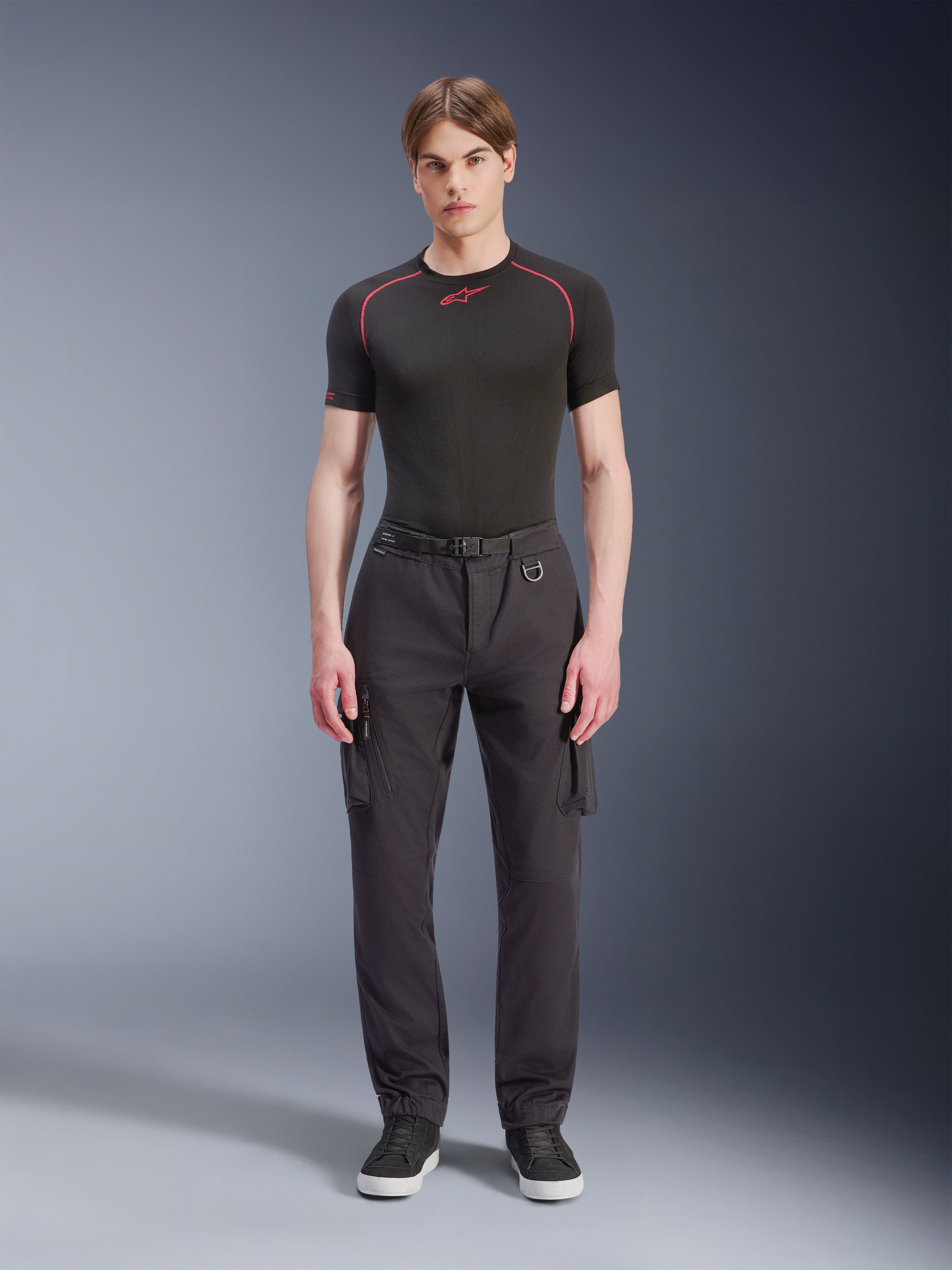 Alpinestars Flex-AST Cargo Hose, Schwarz, urbane Motorradhose, getragen von einem männlichen Model mit schwarzem kurzärmeligem Oberteil und Sneakern, Regular Tapered Passform, Cargotaschen und integrierte Gürtelschnalle