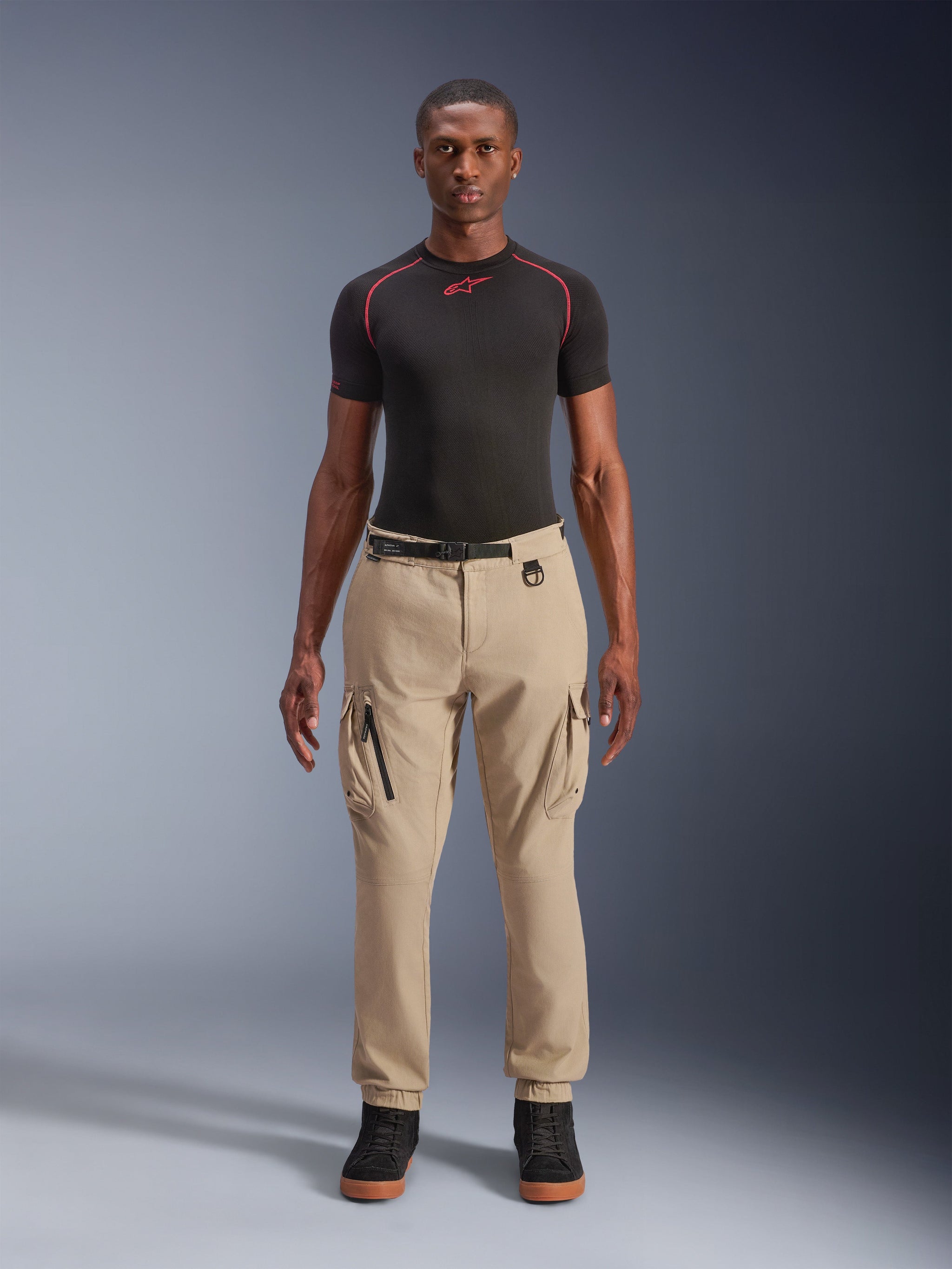 Alpinestars Flex-AST Cargo Pants, Urban Motorcycle Pants, Light Beige, worn by a model with a black technical t-shirt and black sneakers, featuring side cargo pockets with zipper detail, elasticated ankle cuffs, and an adjustable black waist belt, utilitarian riding apparel