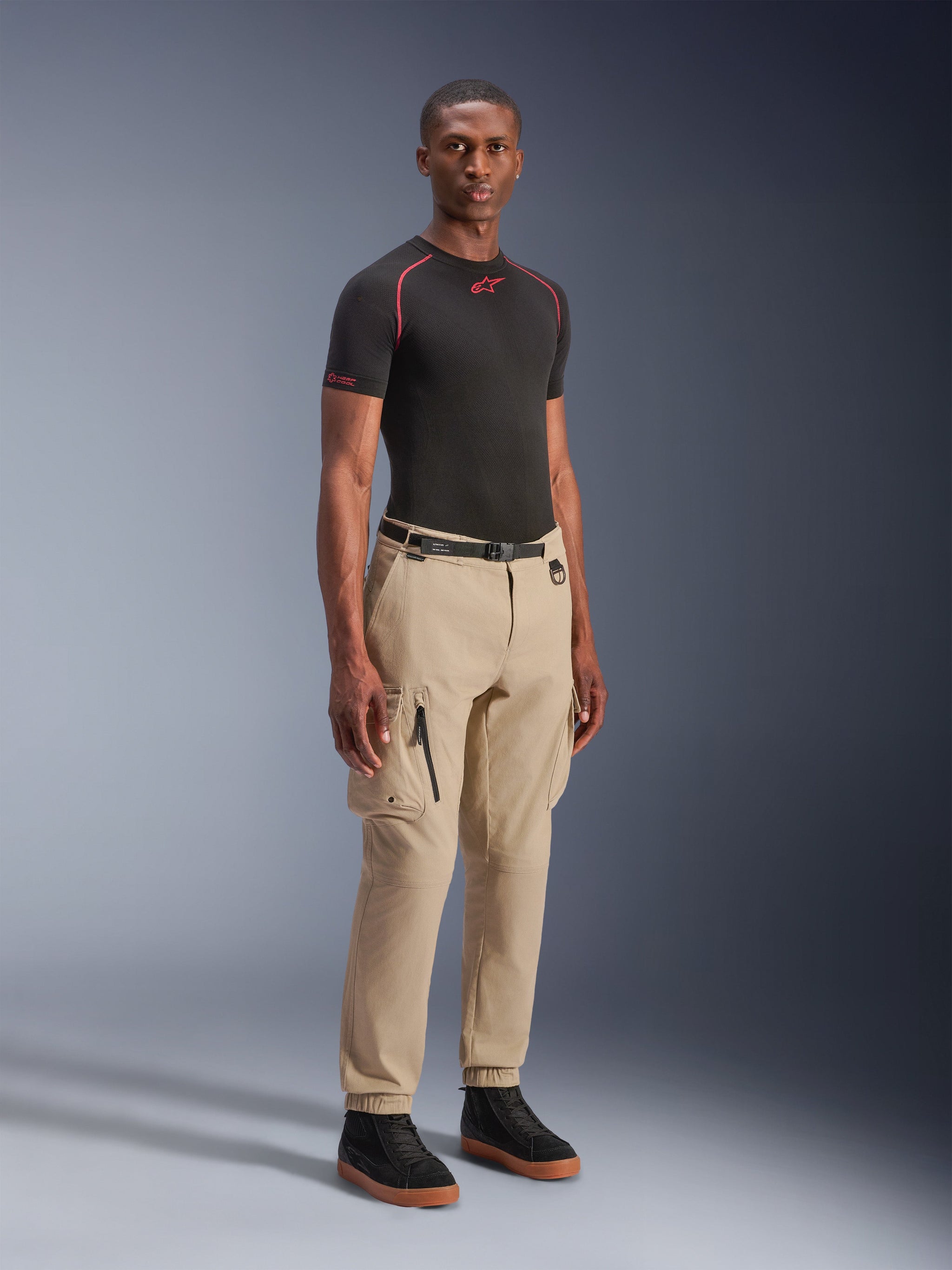 Model wearing Alpinestars Flex-AST Cargo Pants in Light Beige, regular tapered fit with elasticated hems, large cargo pockets on thighs, black waist belt, paired with a black short-sleeve base layer and black sneakers, urban motorcycle style