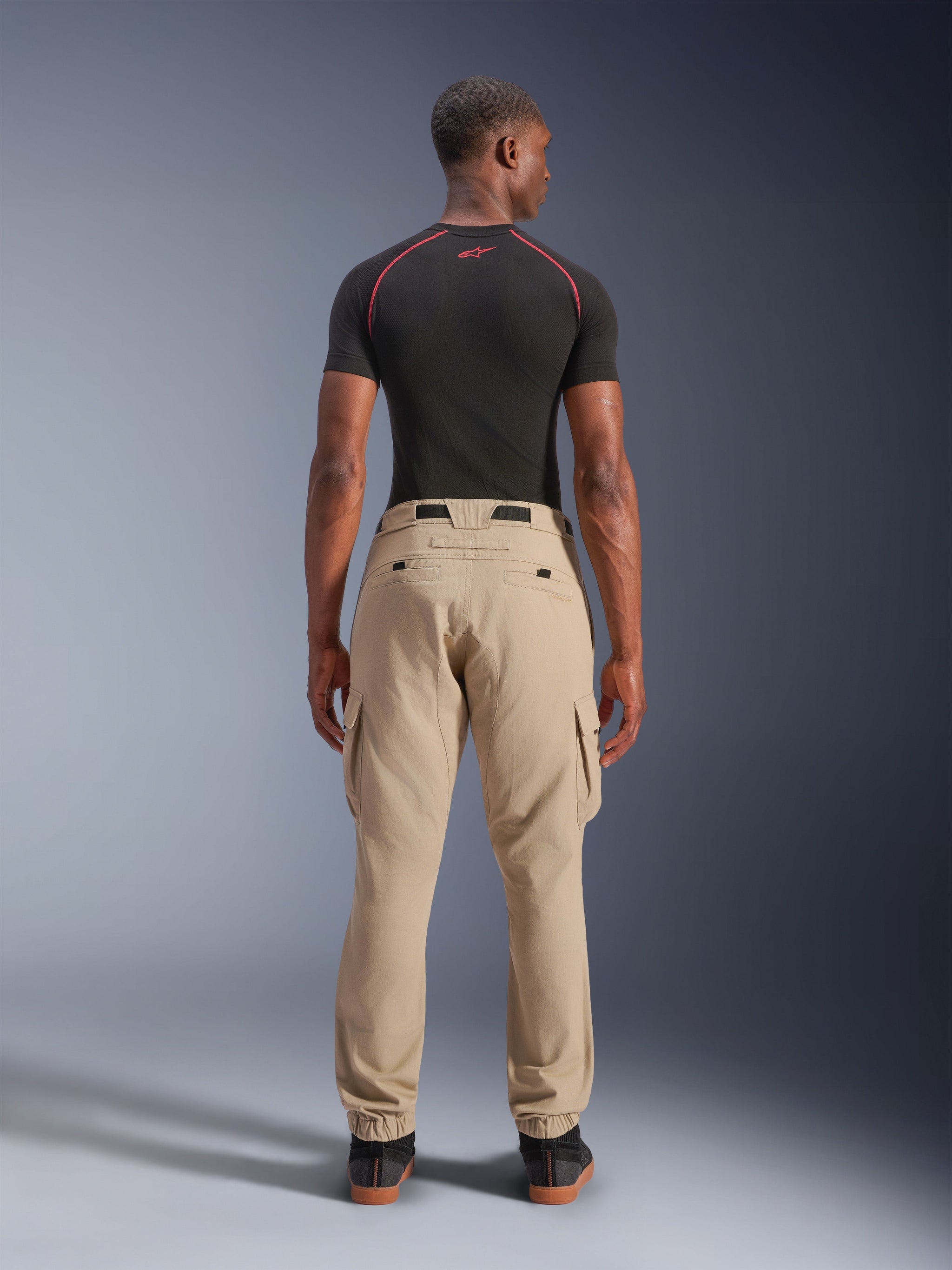 Alpinestars Flex-AST Cargo Pants, Light Beige, back view on a model, regular tapered fit with side cargo pockets and elasticated hems, paired with a black short-sleeve base layer featuring red accents, urban motorcycle gear