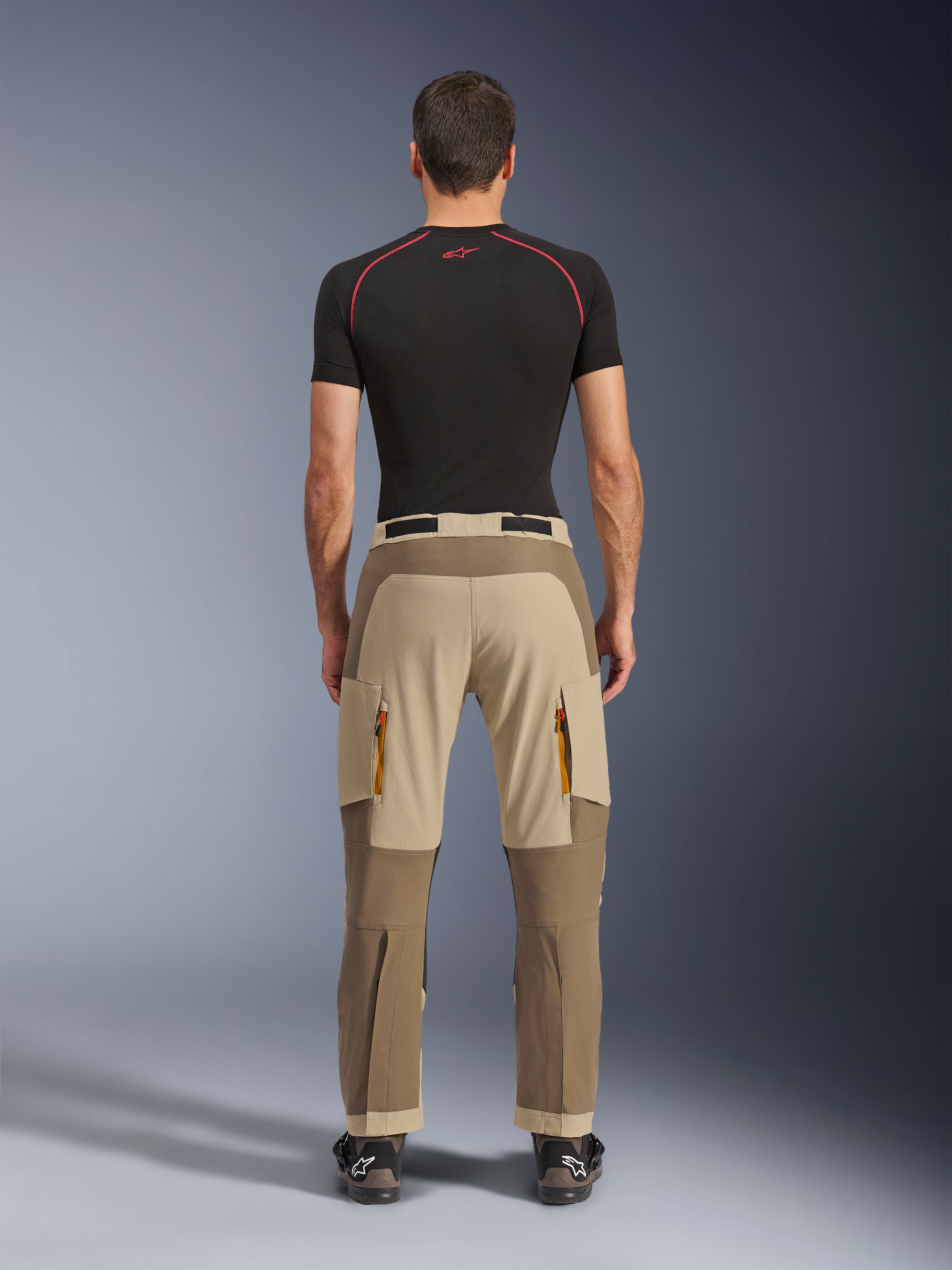 Alpinestars Maxdura Dual Hose in Walnut Aluminum, Rückansicht mit zweifarbigem Design in Braun und Tan, Adventure-Motorradhose mit Reißverschluss-Cargotaschen und verstärktem Gesäßbereich, getragen mit einem schwarzen Kompressionsshirt und Motorradstiefeln.