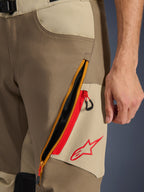 Alpinestars Maxdura Dual Hose, Walnuss und Aluminium, Nahaufnahme des Oberschenkel-Details mit großem diagonalen Reißverschluss-Belüftungssystem, rote Akzente und rotes Alpinestars-Logo auf beigem Einsatz, Adventure-Motorradausrüstung
