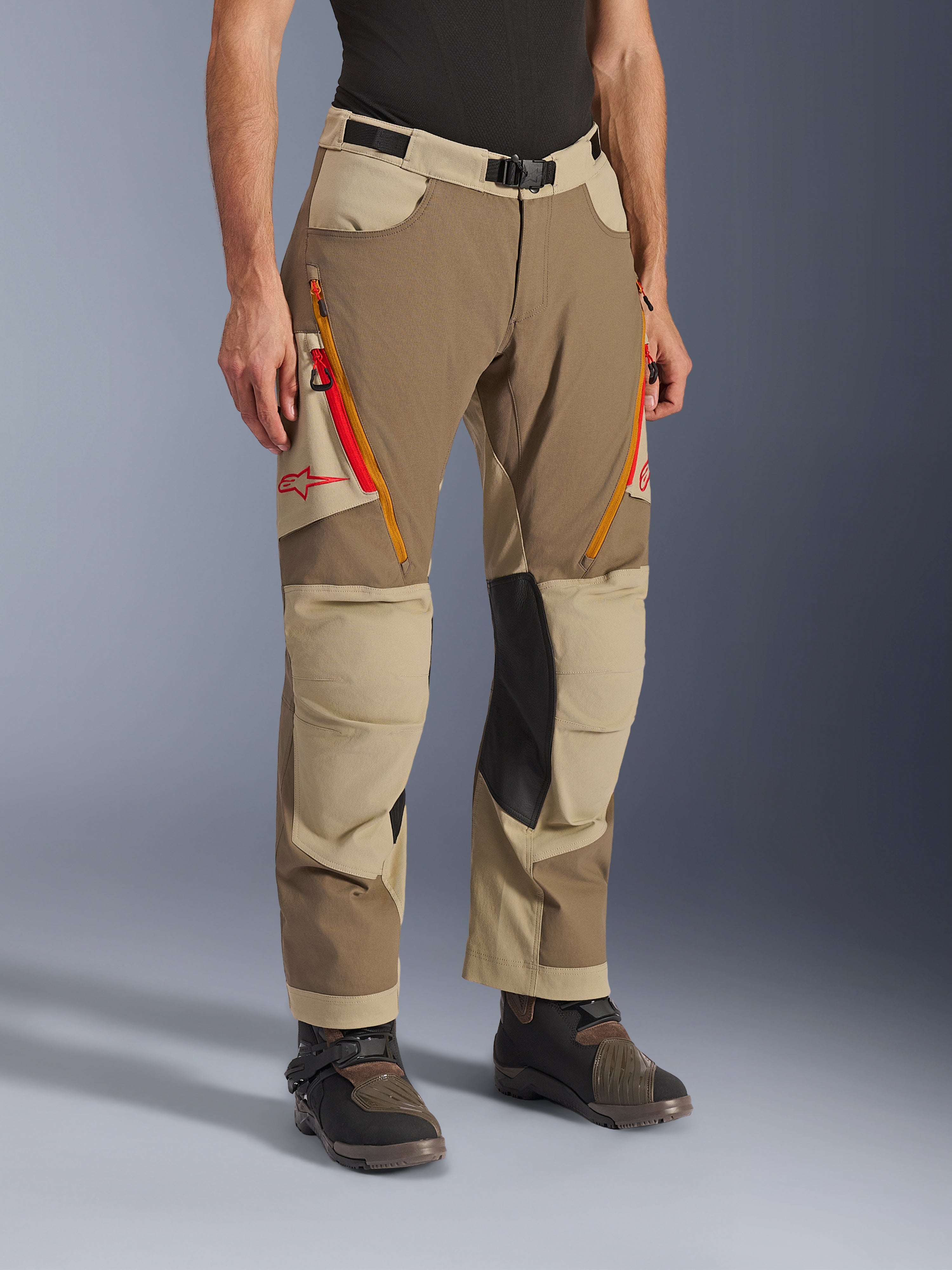 Alpinestars Maxdura Dual Hose, walnut-aluminum-braune und khakifarbene Adventure-Fahrhose, mit orangefarbenen und roten Belüftungsreißverschlüssen an den Oberschenkeln, verstärkten schwarzen Leder-Innenknie-Einsätzen und rotem Astars-Logo, getragen mit technischen Stiefeln.