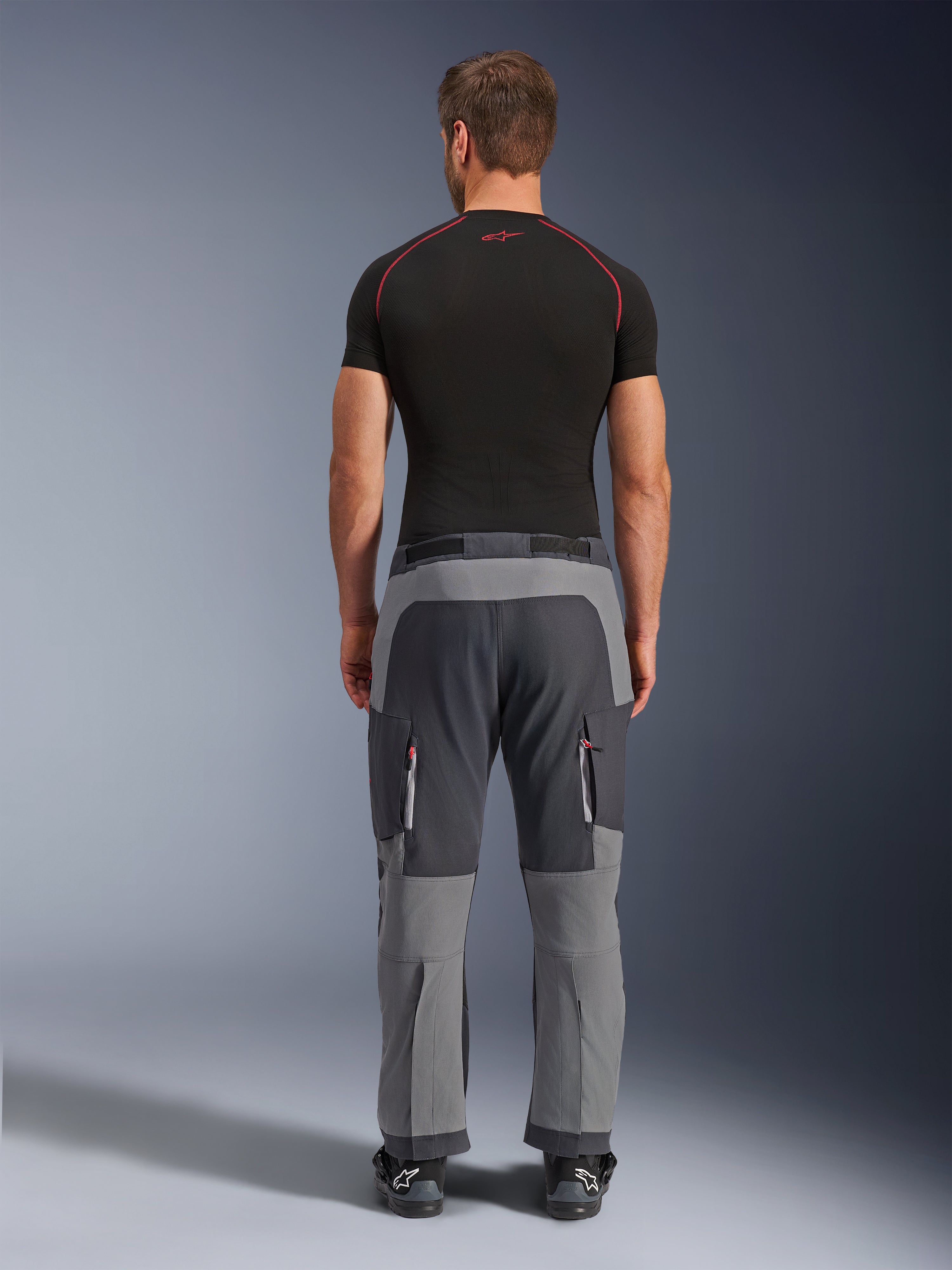 Alpinestars Maxdura Dual Hose, Adventure-Motorradhose in Dunkelgrau und Deep Gray, Rückansicht am Model in schwarzem T-Shirt und Stiefeln, mit mehrfarbigen grauen Einsätzen und Reißverschluss-Cargotaschen mit roten Akzenten.