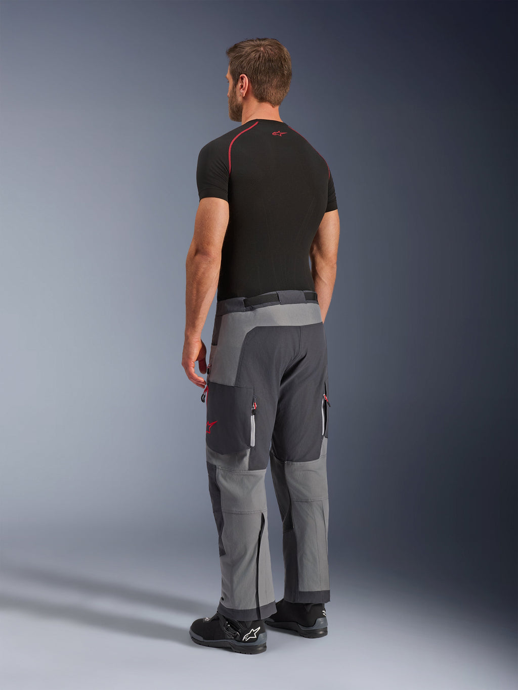 Alpinestars Maxdura Dual Hose, Adventure-Motorradhose, Dunkelgrau und Tiefgrau, Rückansicht am Modell, rote Akzente an Reißverschlüssen und Logo, verstärkte graue Paneele, Cargotaschen und Over-Boot-Passform