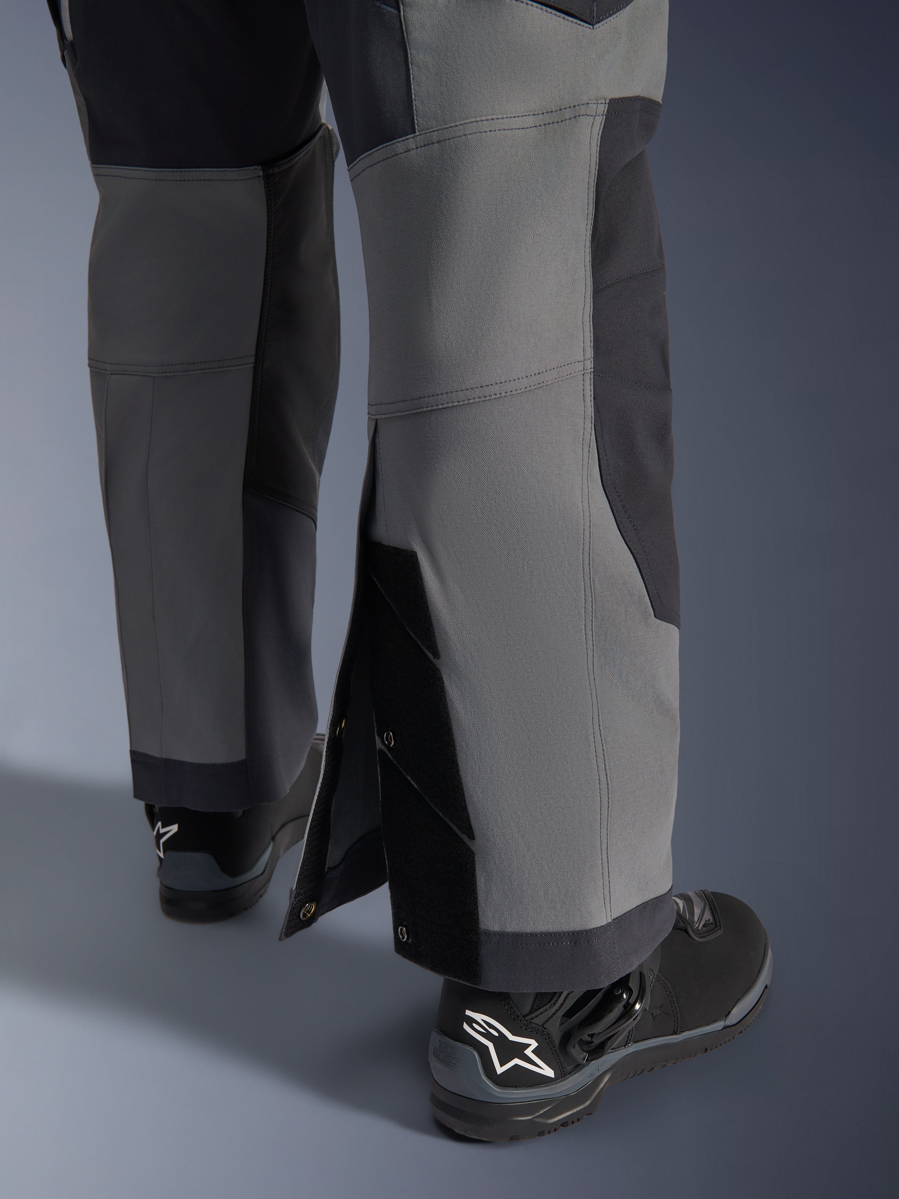 Alpinestars Maxdura Dual Hose, Adventure-Motorradhose, Dunkelgrau und Deep Gray, Nahaufnahme des verstellbaren Over-the-Boot-Beinsaums mit Druckknopfverschlüssen und verstärkten Einsätzen, getragen mit schwarzen Alpinestars Stiefeln, robustes technisches Design für Dual-Sport-Fahrten