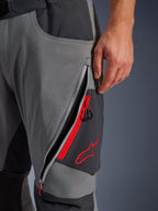 Alpinestars Maxdura Dual Hose, Adventure-Motorradhose in Dunkelgrau und Tiefgrau, Nahaufnahme einer offenen Oberschenkel-Belüftungsöffnung mit Reißverschluss, roten Highlights und gesticktem roten Stern-Logo, technisches Stretch-Gewebe-Design