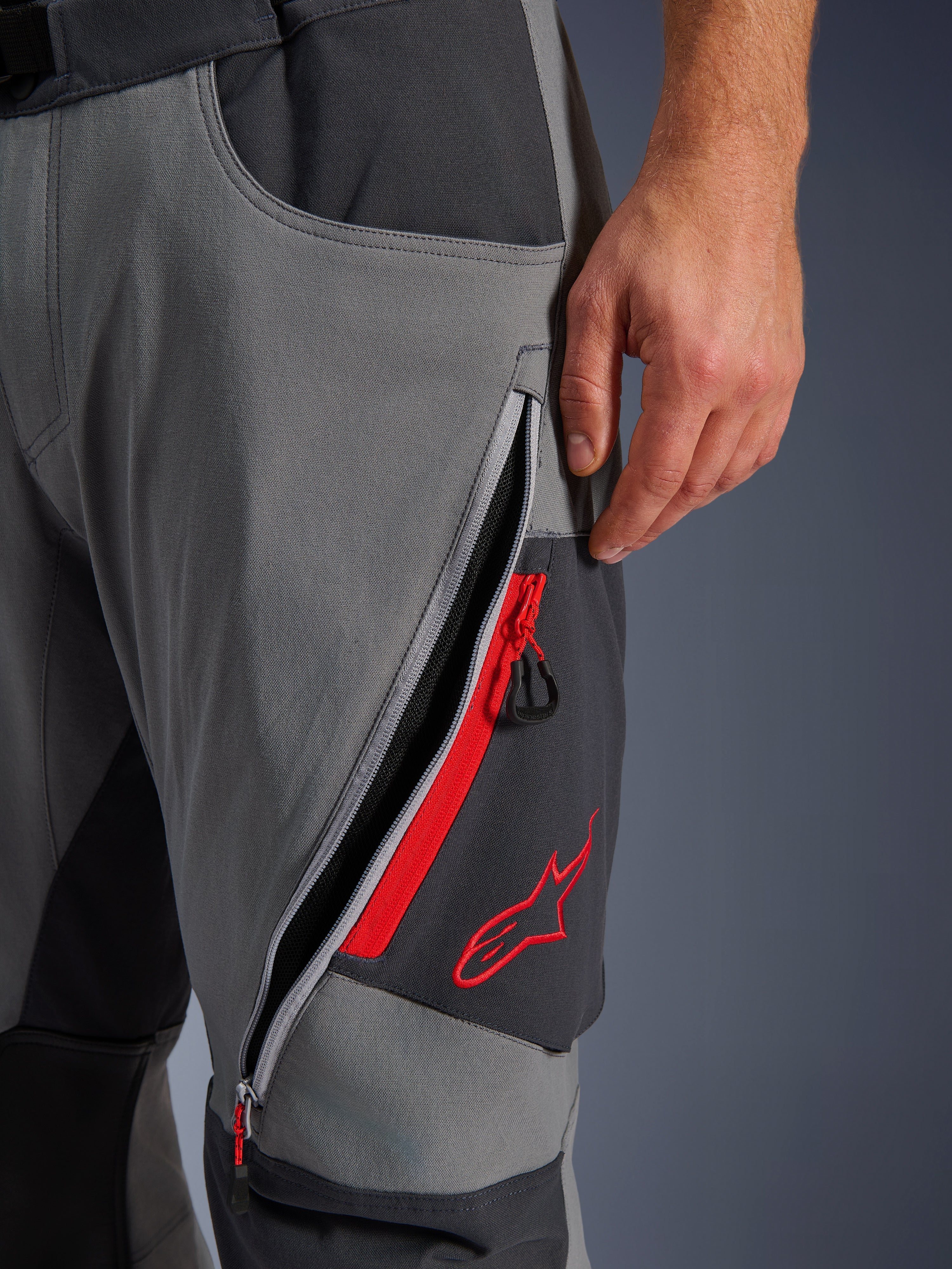 Alpinestars Maxdura Dual Hose, Adventure-Motorradhose in Dunkelgrau und Tiefgrau, Nahaufnahme einer offenen Oberschenkel-Belüftungsöffnung mit Reißverschluss, roten Highlights und gesticktem roten Stern-Logo, technisches Stretch-Gewebe-Design