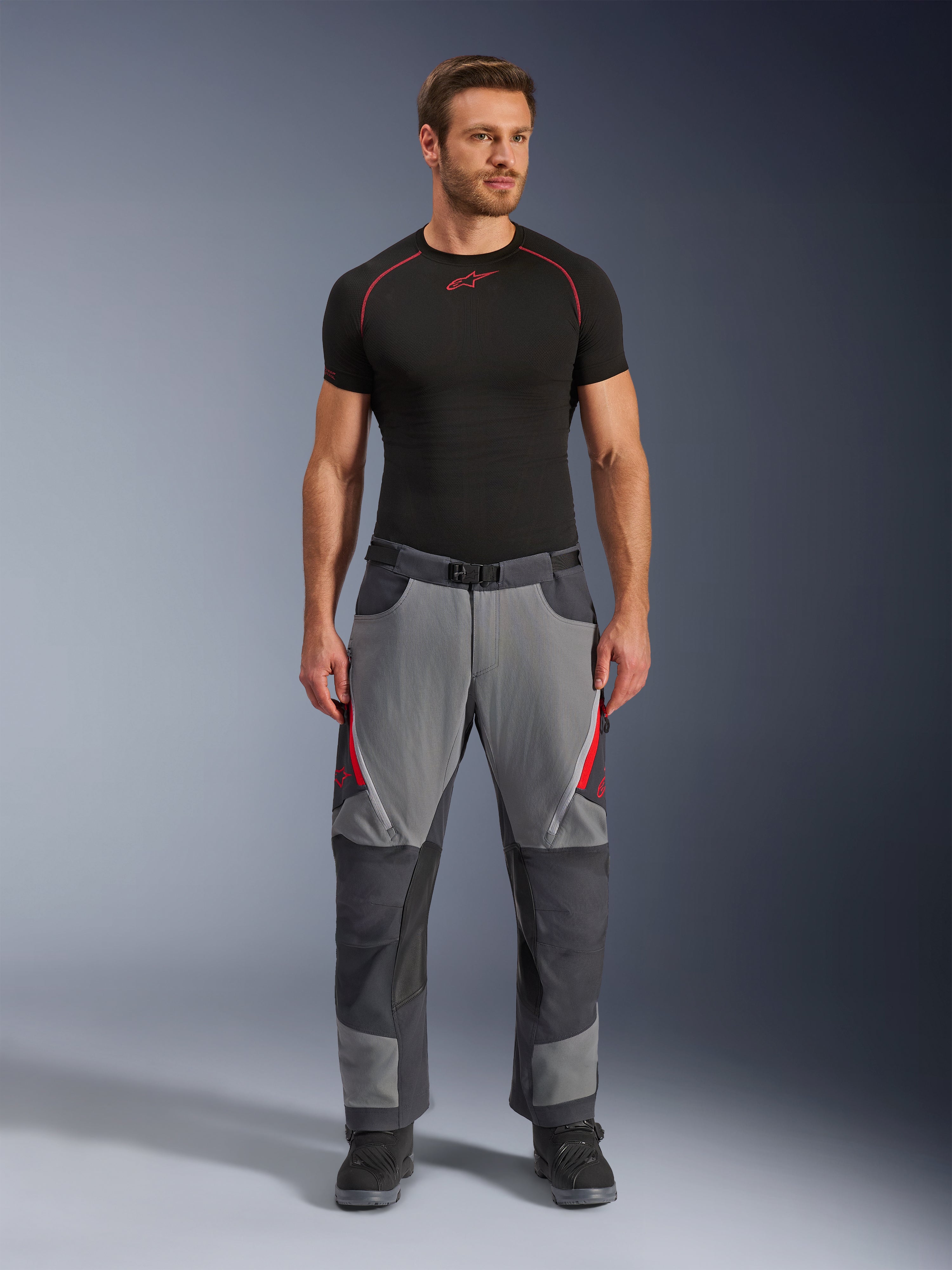 Alpinestars Maxdura Dual Hose, Motorradhose, Dunkelgrau und Tiefgrau mit roten Akzenten, Vorderansicht getragen von einem Model mit schwarzem Alpinestars Base Layer und schwarzen Stiefeln, mit verstärkten Paneelen und Oberschenkel-Belüftungsöffnungen mit Reißverschluss, Adventure-Stil Fahrausrüstung