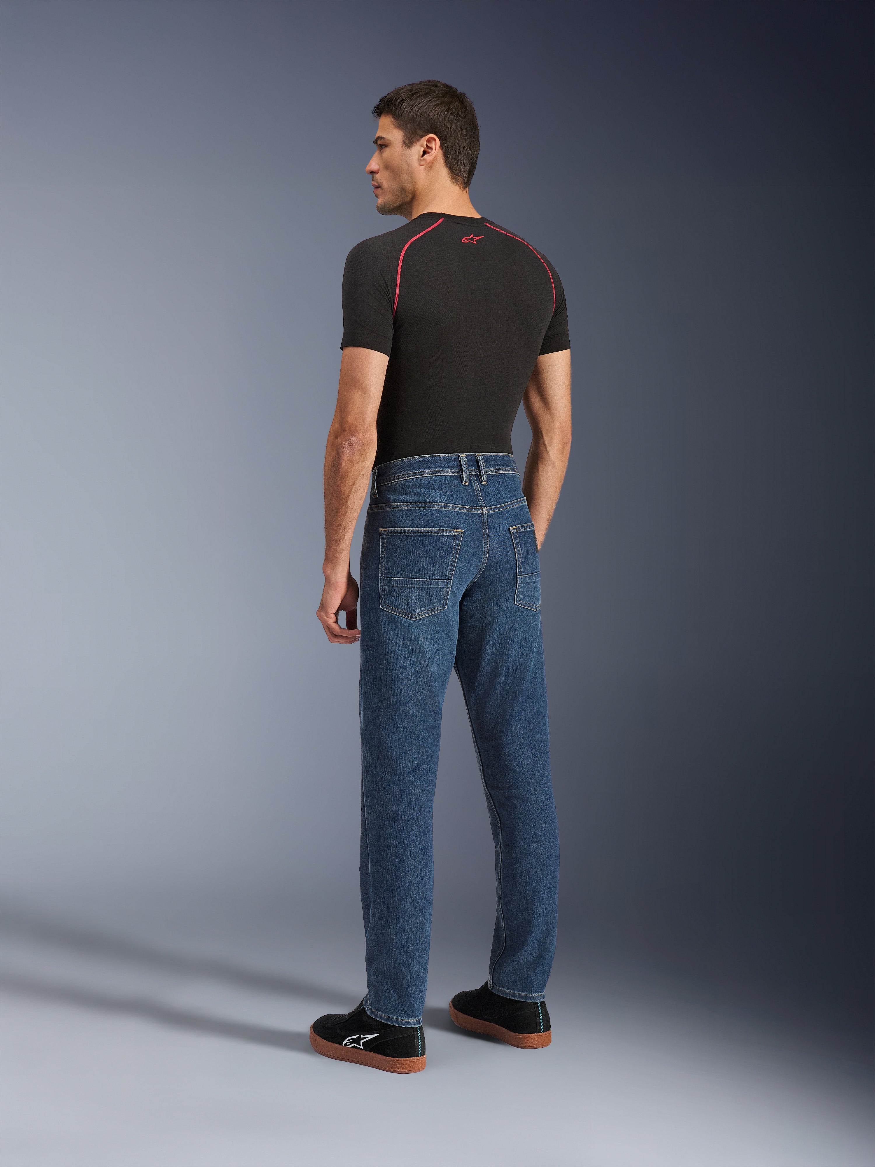 Alpinestars Flex-AST 5 Denim-Hose, Indigo Washed, urbane Motorradhose mit konisch zulaufender Passform, Rückansicht an einem Modell in schwarzem T-Shirt und Sneakern, mit schlichtem Five-Pocket-Design und Monolayer-Stretch-Konstruktion