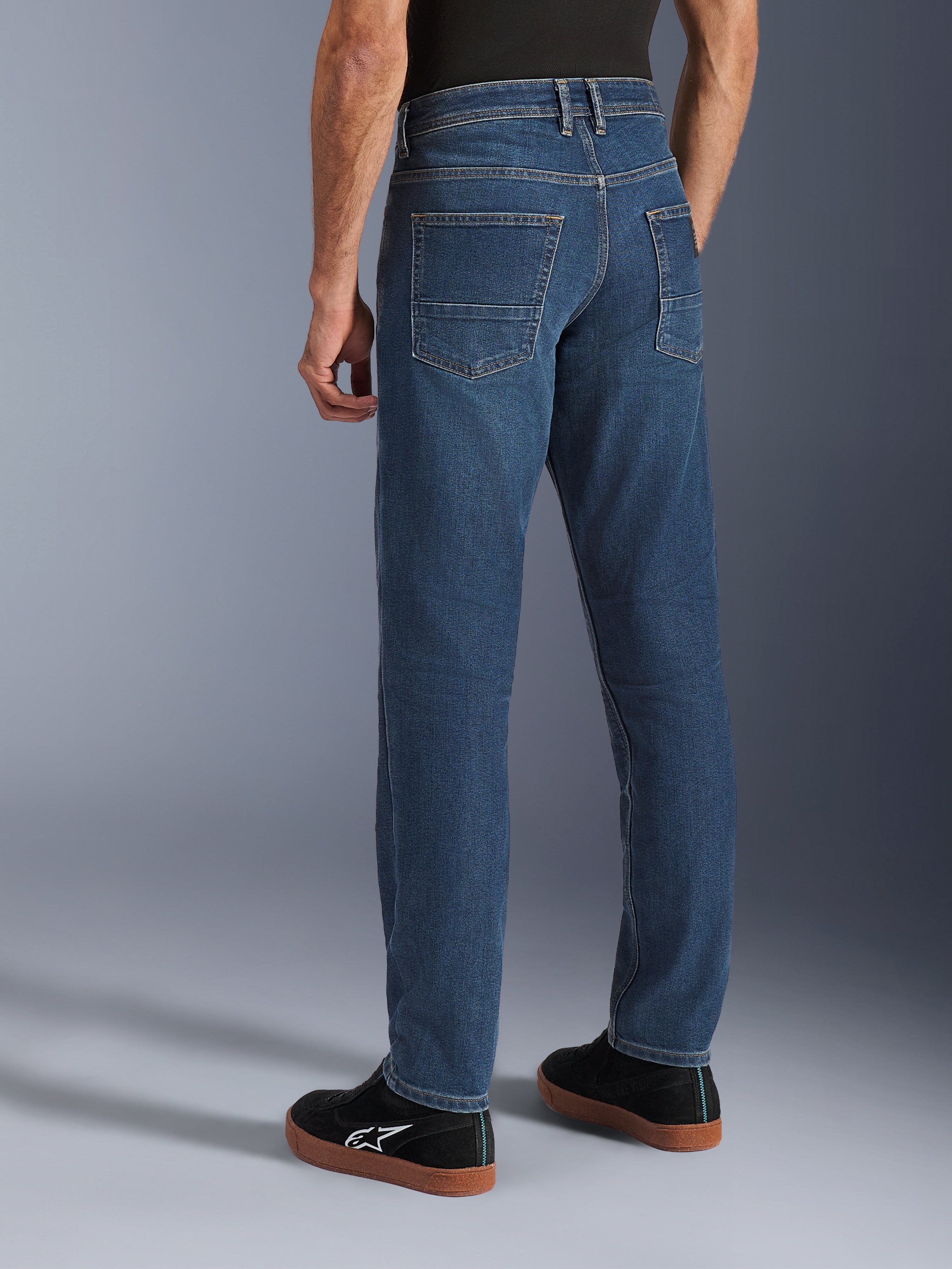 Alpinestars Flex-AST 5 Denim-Hose, Indigo Washed Blau, Rückansicht einer Person in lässigen Motorradjeans mit konischer Passform, kombiniert mit schwarzen Alpinestars-Sneakern, mit Gesäßtaschen und verstärkten Nähten für urbane Fahrten