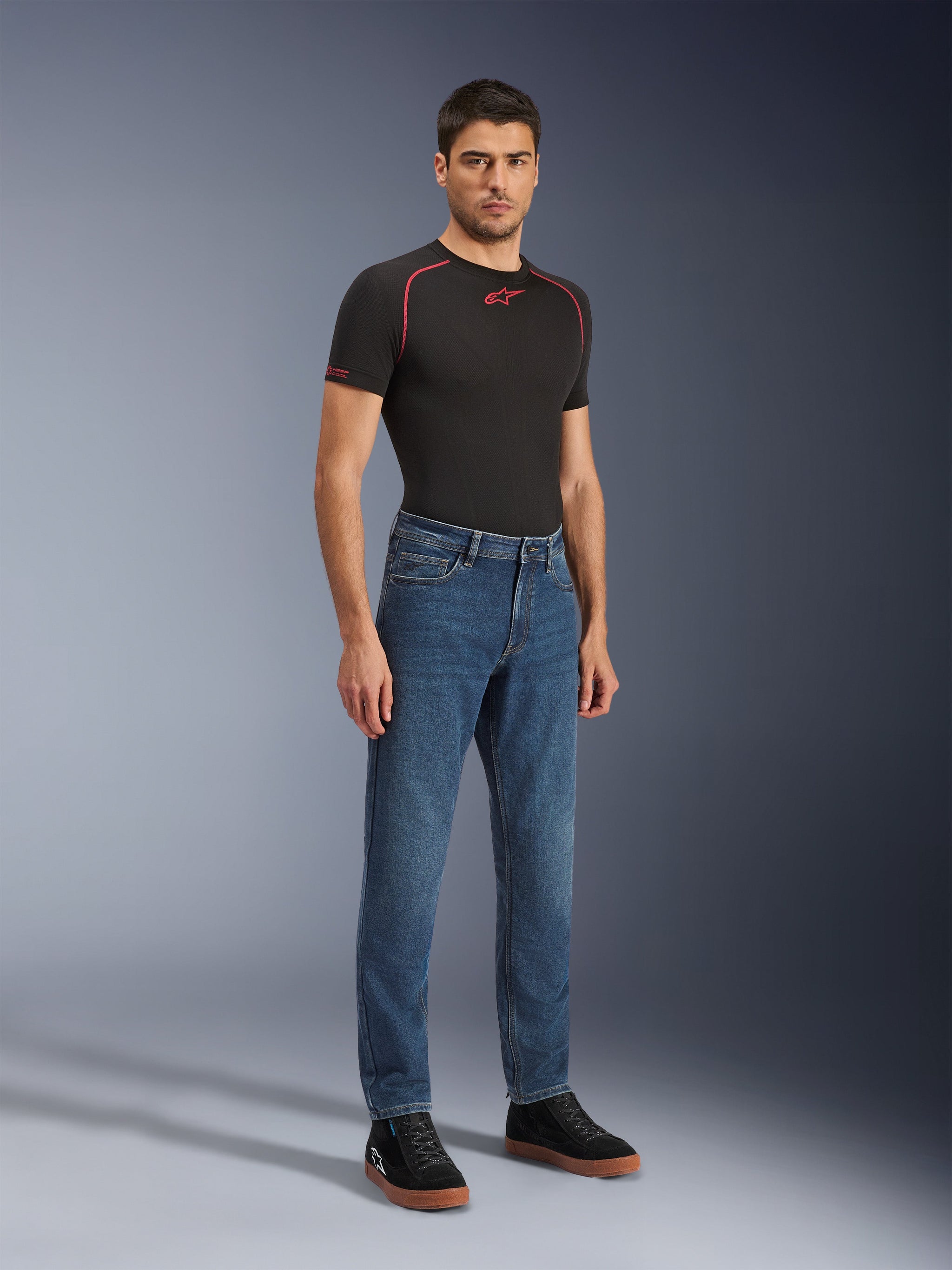 Alpinestars Flex-AST 5 Denim Hose - Short, Indigo Washed, urbane Motorradjeans mit konisch zulaufender Passform, getragen von einem männlichen Model mit schwarzem, eng anliegendem Shirt und schwarzen Sneakern, klassischer Denim-Stil für den Übergang von der Straße zum Alltag