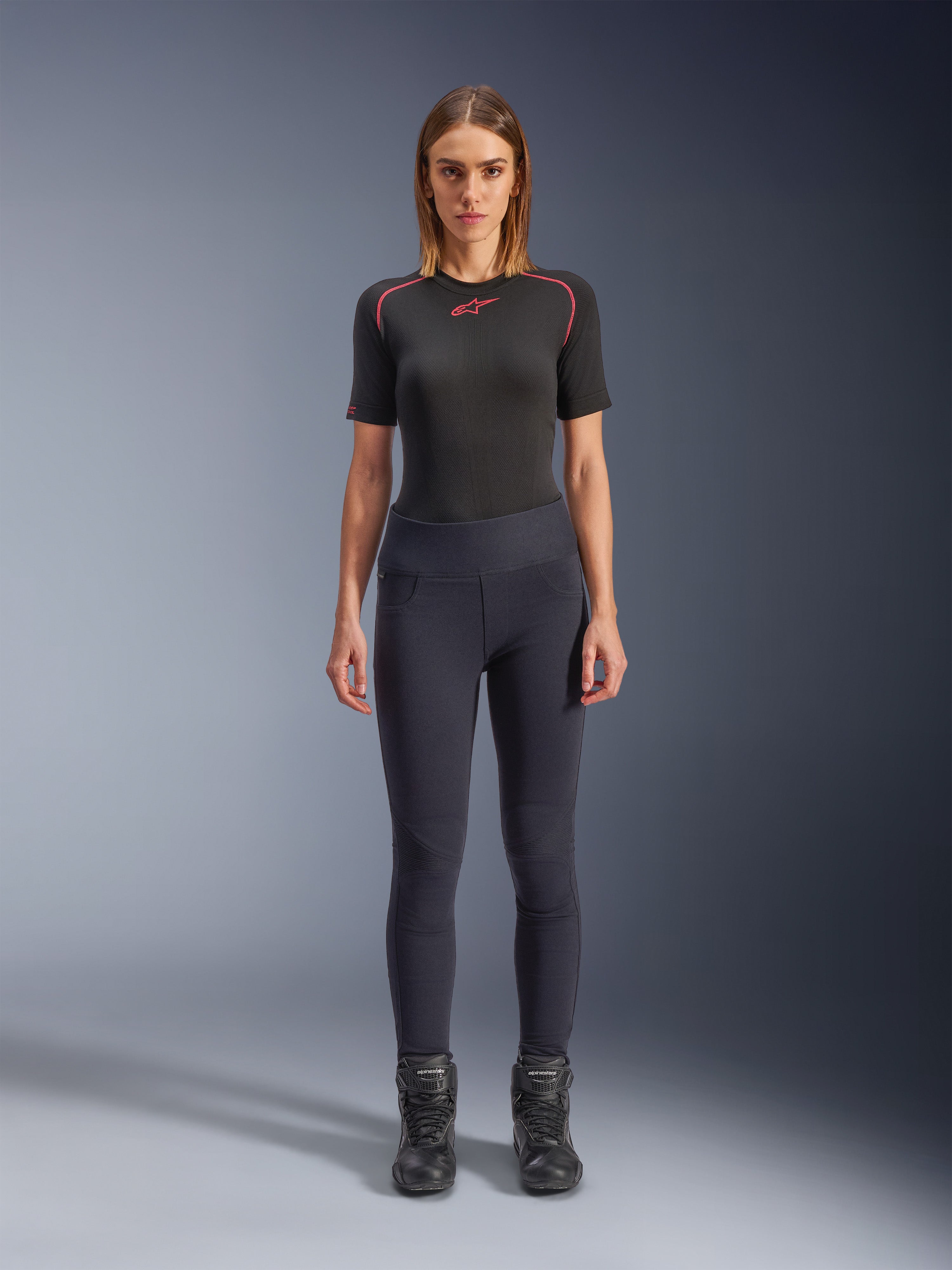 Alpinestars Stella Flex-AST Leggings, urbane Damen-Motorrad-Leggings in Schwarz, schmal geschnittenes High-Waist-Design, getragen mit einem schwarzen, kurzärmeligen Funktions-Oberteil und schwarzen Motorradstiefeln, mit Akkordeon-Kniepaneelen und einem modernen, minimalistischen Look.
