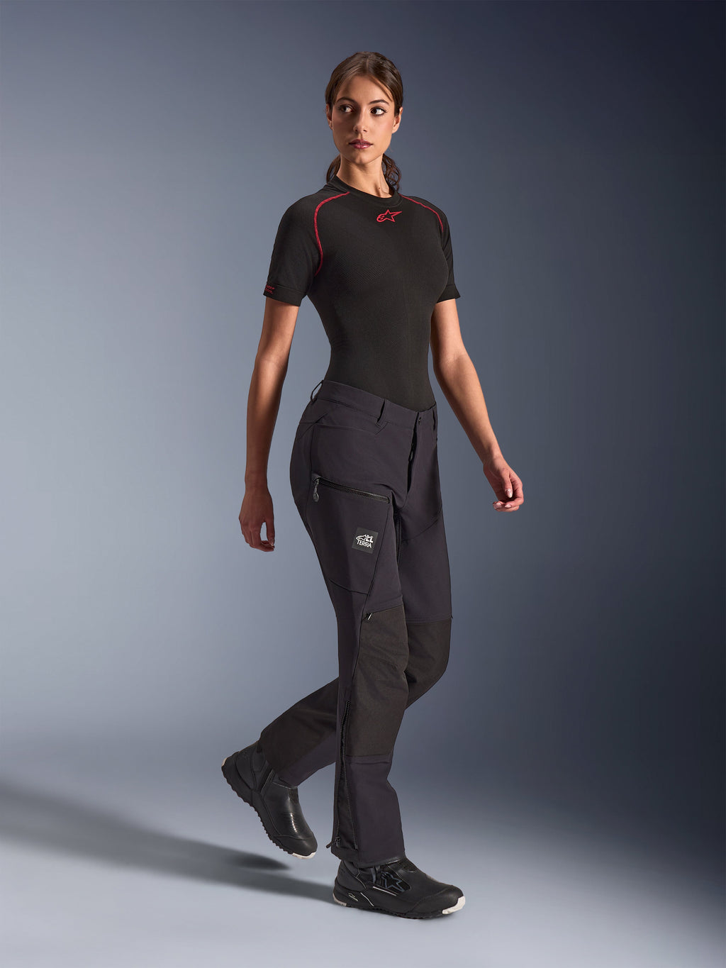 Alpinestars Stella Flex-AST Explorer Damen-Hose, schwarze Adventure-Motorradhose, Stretch-Textilkonstruktion mit Ripstop-Verstärkungen, Cargo-Taschen mit Reißverschluss und Reißverschlüsse an den Unterschenkeln, getragen mit einem schwarzen kurzärmeligen Base-Layer und Motorradschuhen.