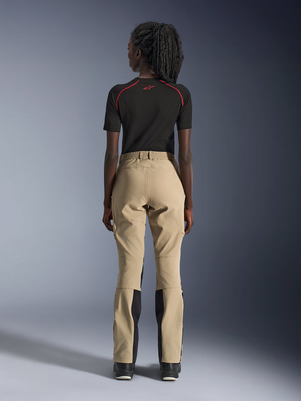 Alpinestars Stella Flex-AST Explorer Hose für Damen in Sand, Rückansicht an einem Model, kombiniert mit einem schwarzen kurzärmeligen Baselayer und schwarzen Stiefeln, mit verstärkten Gesäßpaneelen und ergonomischer Textilkonstruktion für Adventure-Touren.