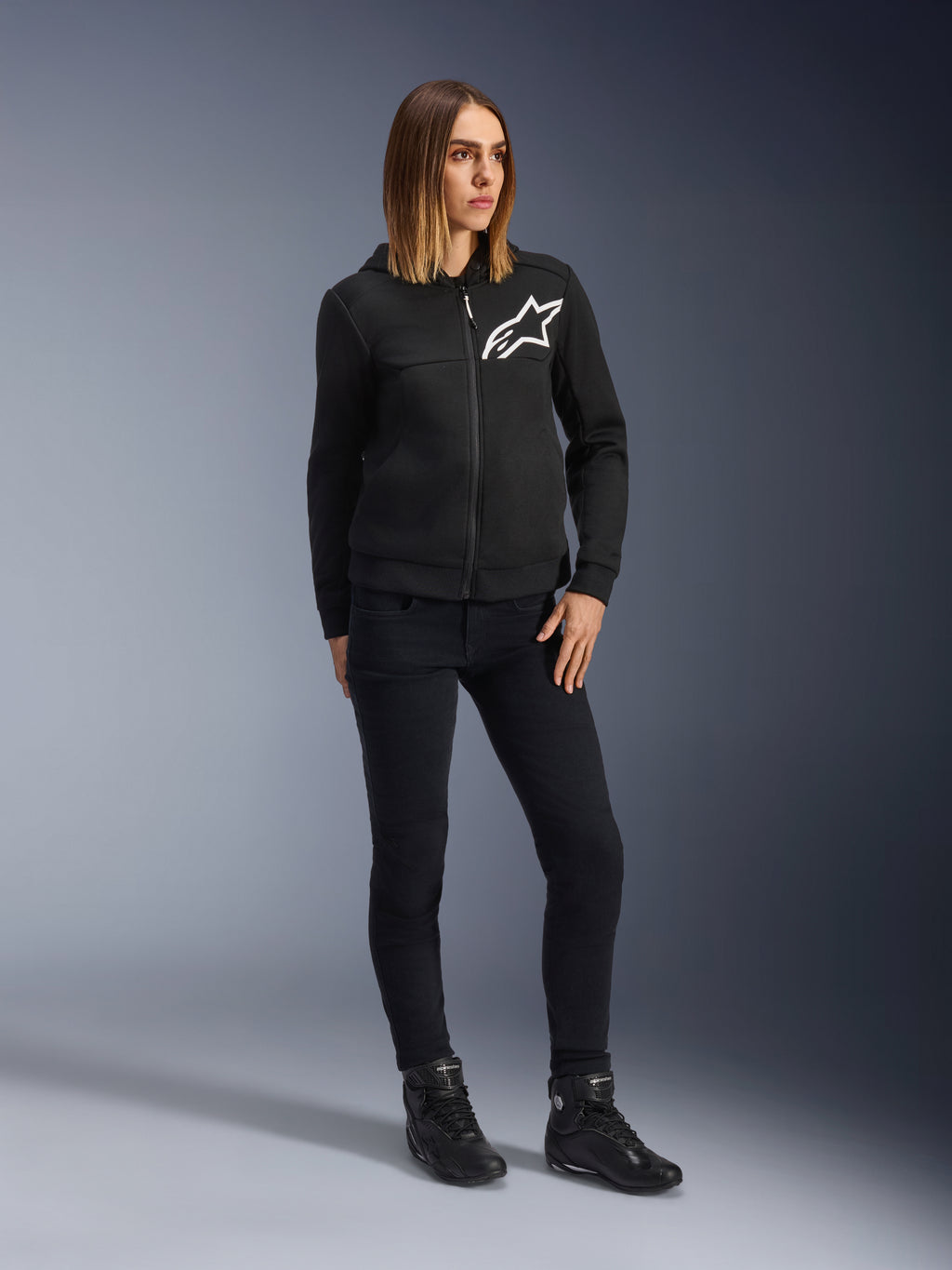 Frau trägt Alpinestars Daisy V3 Damen-Motorradjeans, schmal geschnittene Motorradhose in Rinse Black, kombiniert mit einer schwarzen Kapuzenjacke und Motorradstiefeln, urbane lässige Schutzausrüstung