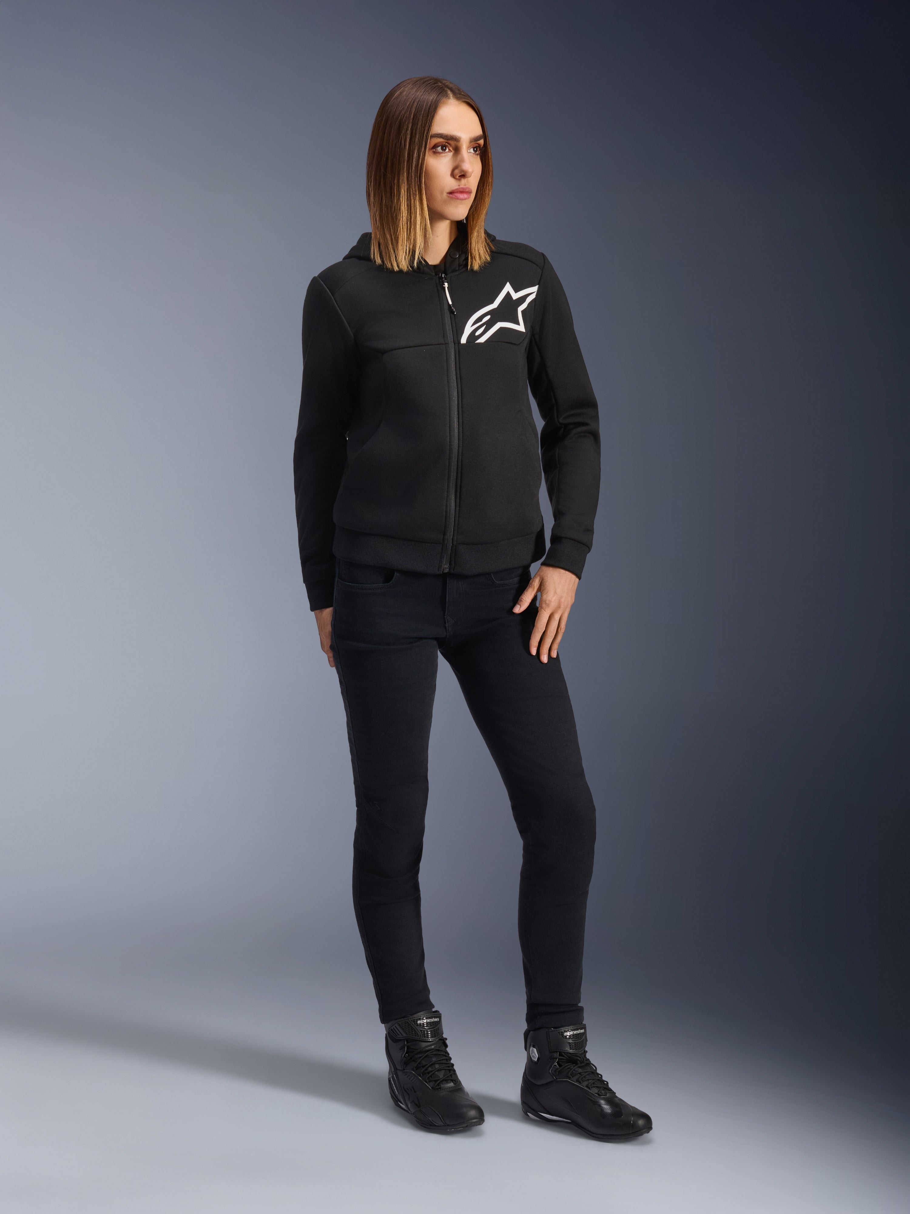 Frau trägt Alpinestars Daisy V3 Damen-Motorradjeans, schmal geschnittene Motorradhose in Rinse Black, kombiniert mit einer schwarzen Kapuzenjacke und Motorradstiefeln, urbane lässige Schutzausrüstung