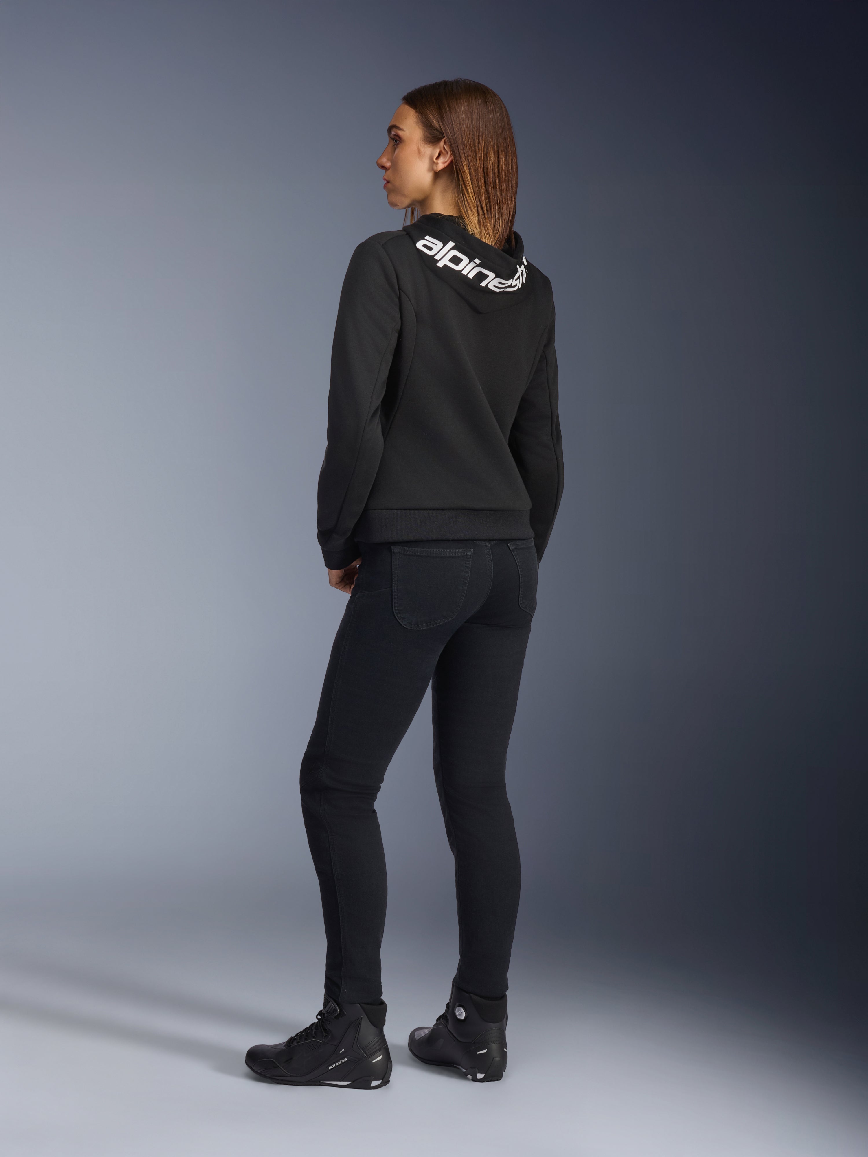 Alpinestars Daisy V3 Damen-Motorradjeans, Damen-Motorradhose, Rinse Black, Rückansicht zeigt Slim-Fit-Passform und hohen Bund hinten, kombiniert mit einem schwarzen Alpinestars-Hoodie und Motorradstiefeln für einen urbanen Motorrad-Look