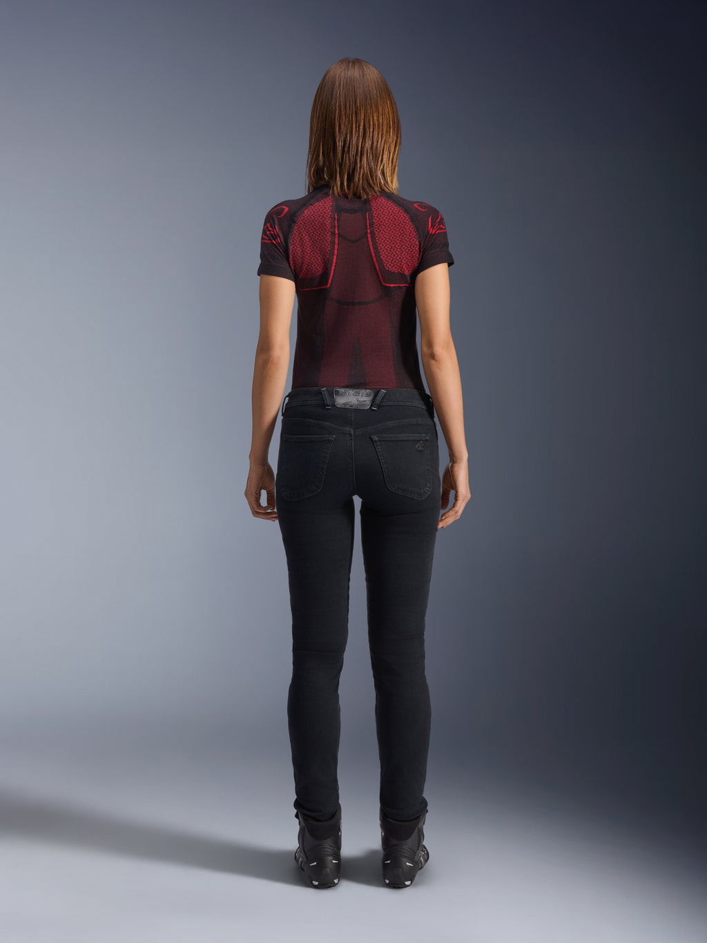 Alpinestars Daisy V3 Damen-Motorradjeans, Rinse Black, schmal geschnittene Motorradhose in der Rückansicht, mit High-Waist-Design und Gesäßtaschen, kombiniert mit einem rot-schwarz gemusterten Funktionsshirt und schwarzen Stiefeln, urbane Motorradausrüstung für Damen