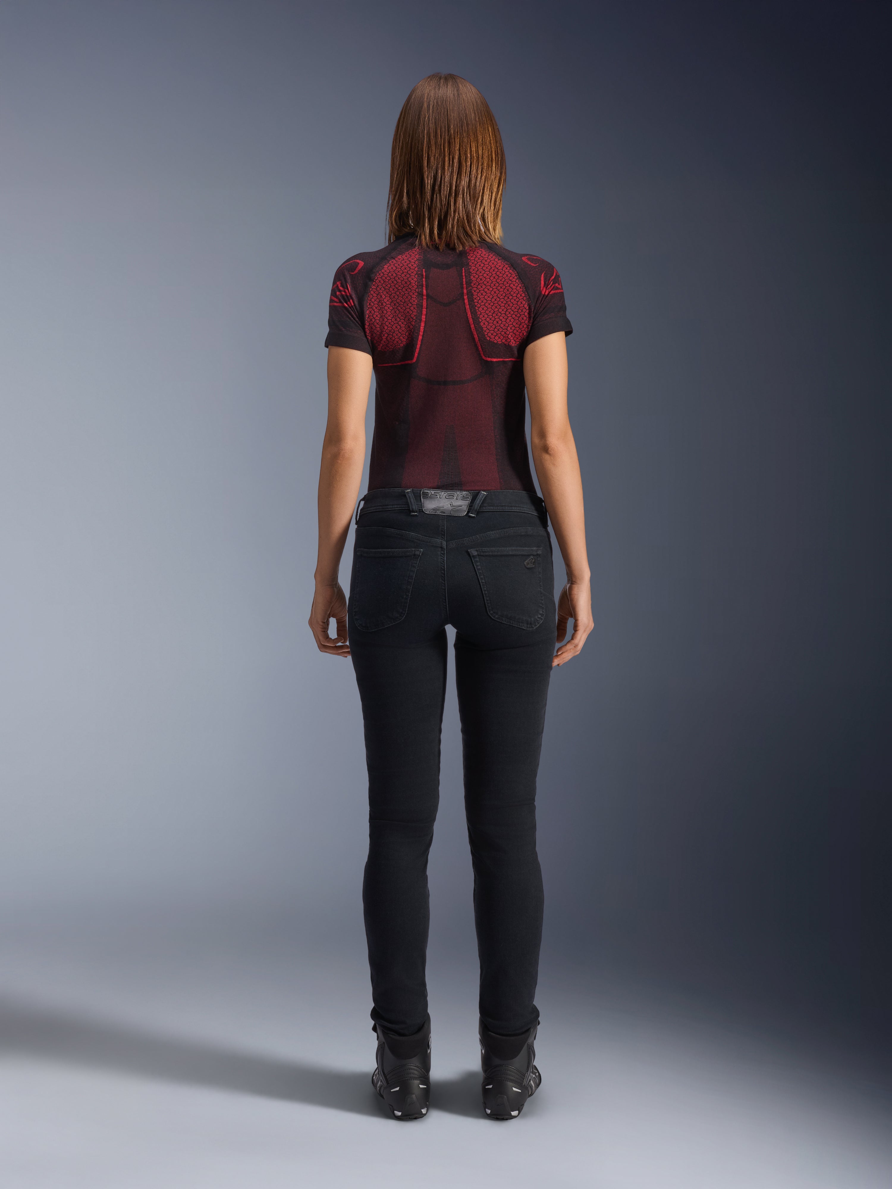 Alpinestars Daisy V3 Damen-Motorradjeans, Rinse Black, schmal geschnittene Motorradhose in der Rückansicht, mit High-Waist-Design und Gesäßtaschen, kombiniert mit einem rot-schwarz gemusterten Funktionsshirt und schwarzen Stiefeln, urbane Motorradausrüstung für Damen