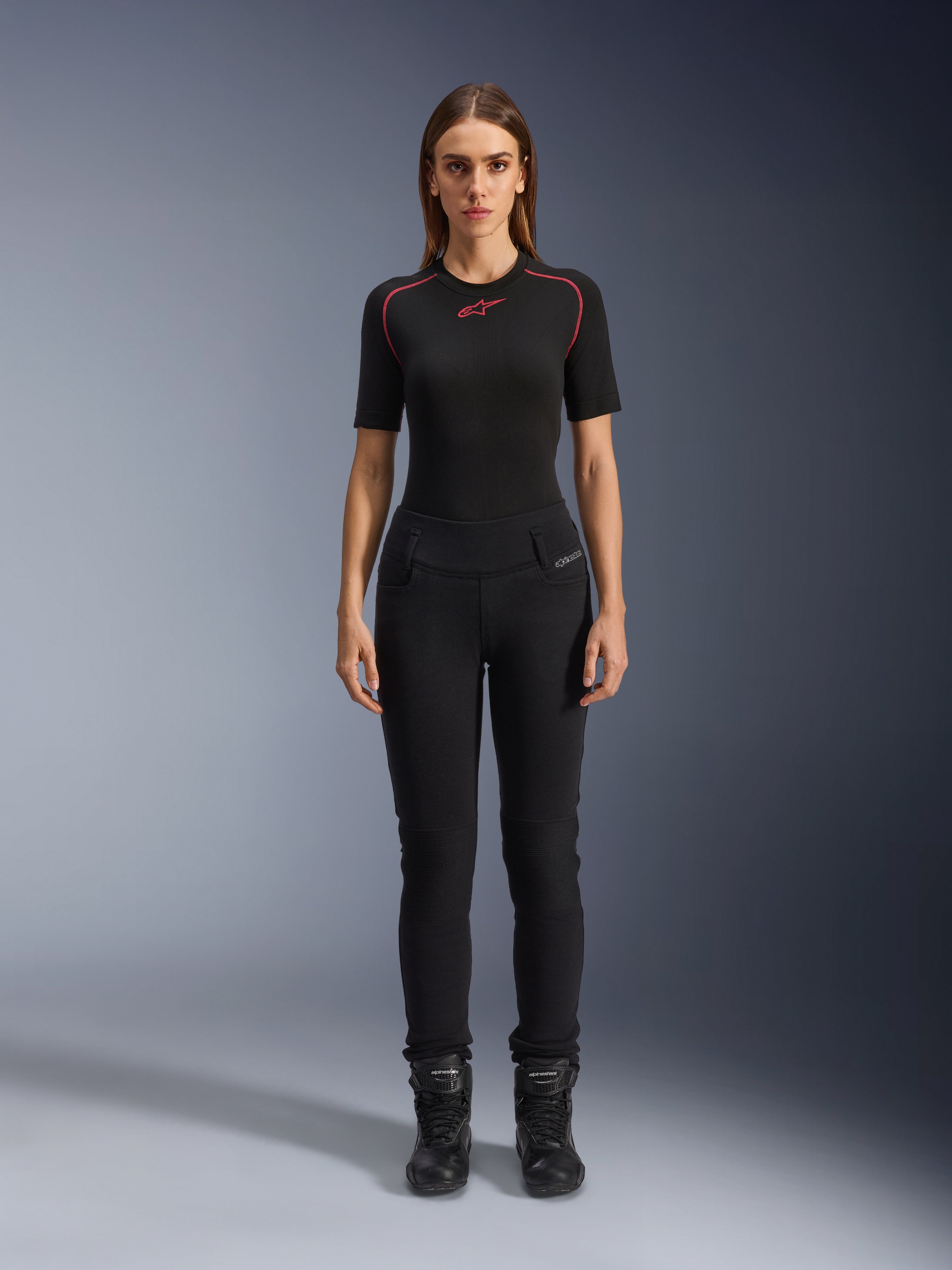 Alpinestars Banshee Damen-Leggings, schwarze Motorrad-Leggings, figurbetonte anatomische Passform, getragen von einem weiblichen Model mit schwarzem Baselayer und Motorradstiefeln, mit gerippten Knieeinsätzen und hoch geschnittenem Bund für urbane Fahrten.