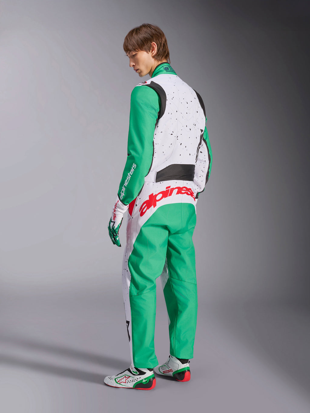 Alpinestars KMX-9 V3 Graphic 6 Anzug, Karting-Anzug, Weiß, Grün und Rot, Modell in der Rückansicht, mit grünen Ärmeln und unteren Einsätzen, weißer oberer Torso mit schwarzen Sprenkel-Details, auffälliges rotes Alpinestars-Branding am unteren Rücken, kombiniert mit passenden weiß-grünen Karting-Schuhen.