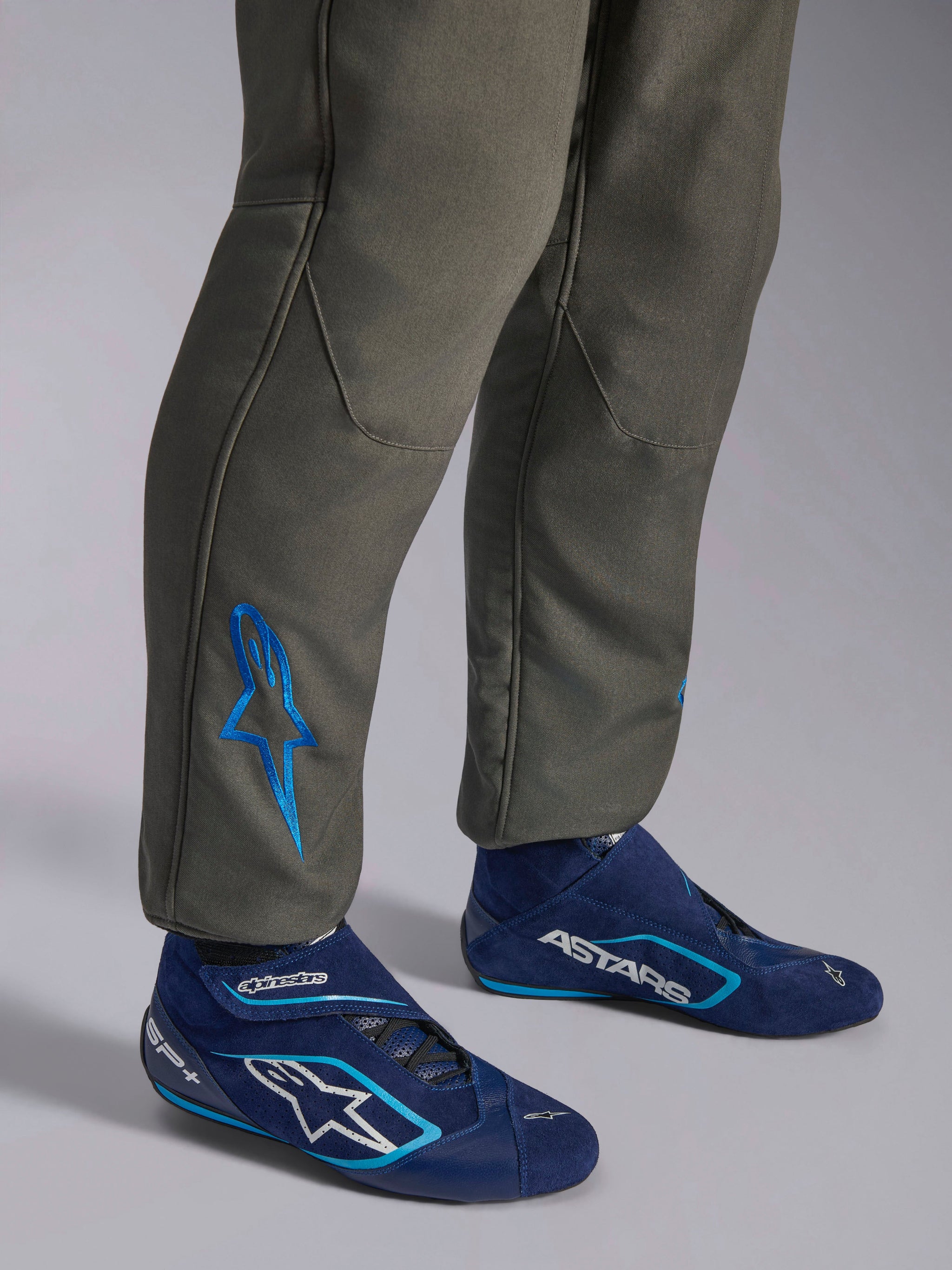 Alpinestars GP Race V3 Anzug FIA, Autorennanzug in Anthrazit-Blau, Nahaufnahme der Unterschenkel mit einem blau bestickten Astars-Logo am Knöchel, kombiniert mit marineblauen und cyanfarbenen Rennstiefeln, professionelle Motorsport-Schutzausrüstung