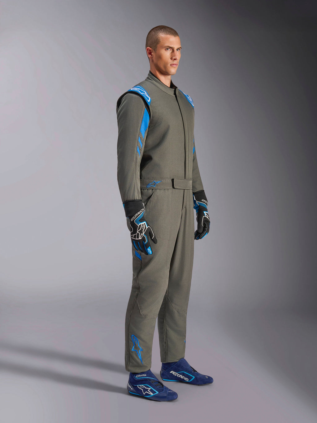 Alpinestars GP Race V3 Anzug FIA in Anthrazit-Blau, einteiliger Auto-Rennanzug getragen von einem männlichen Model, mit leuchtend blauen Akzenten an den Schultern und Knöcheln, kombiniert mit passenden Rennhandschuhen und blauen Rennschuhen, professionelle Motorsport-Ausrüstung.