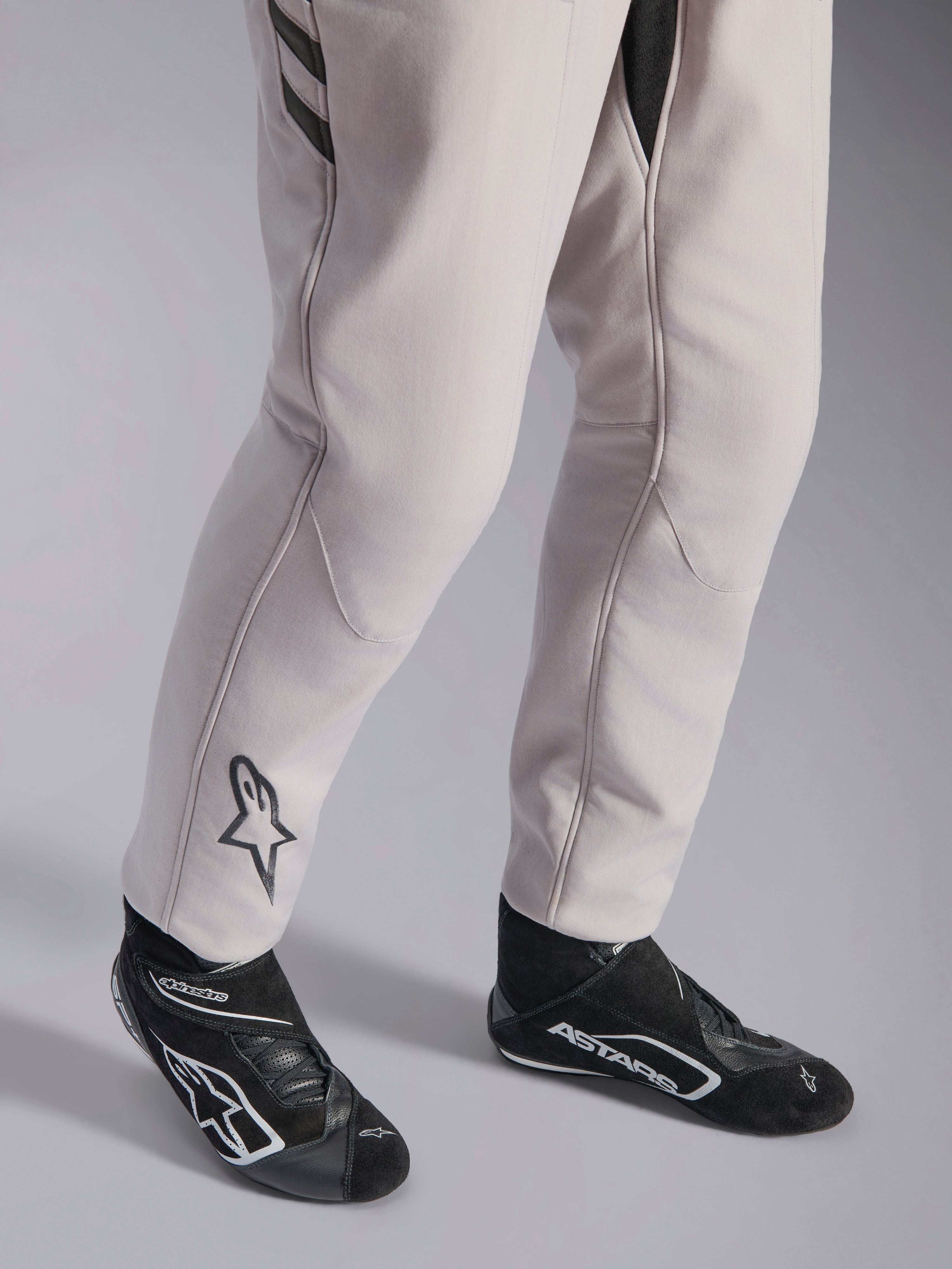Alpinestars GP Race V3 Anzug FIA, Automobil-Rennanzug in Silver Anthracite, Detailaufnahme der Unterschenkel und schwarzen Rennstiefel, mit dem schwarzen Astars-Logo auf der Wade, professionelle Motorsportausrüstung mit Rippbündchen