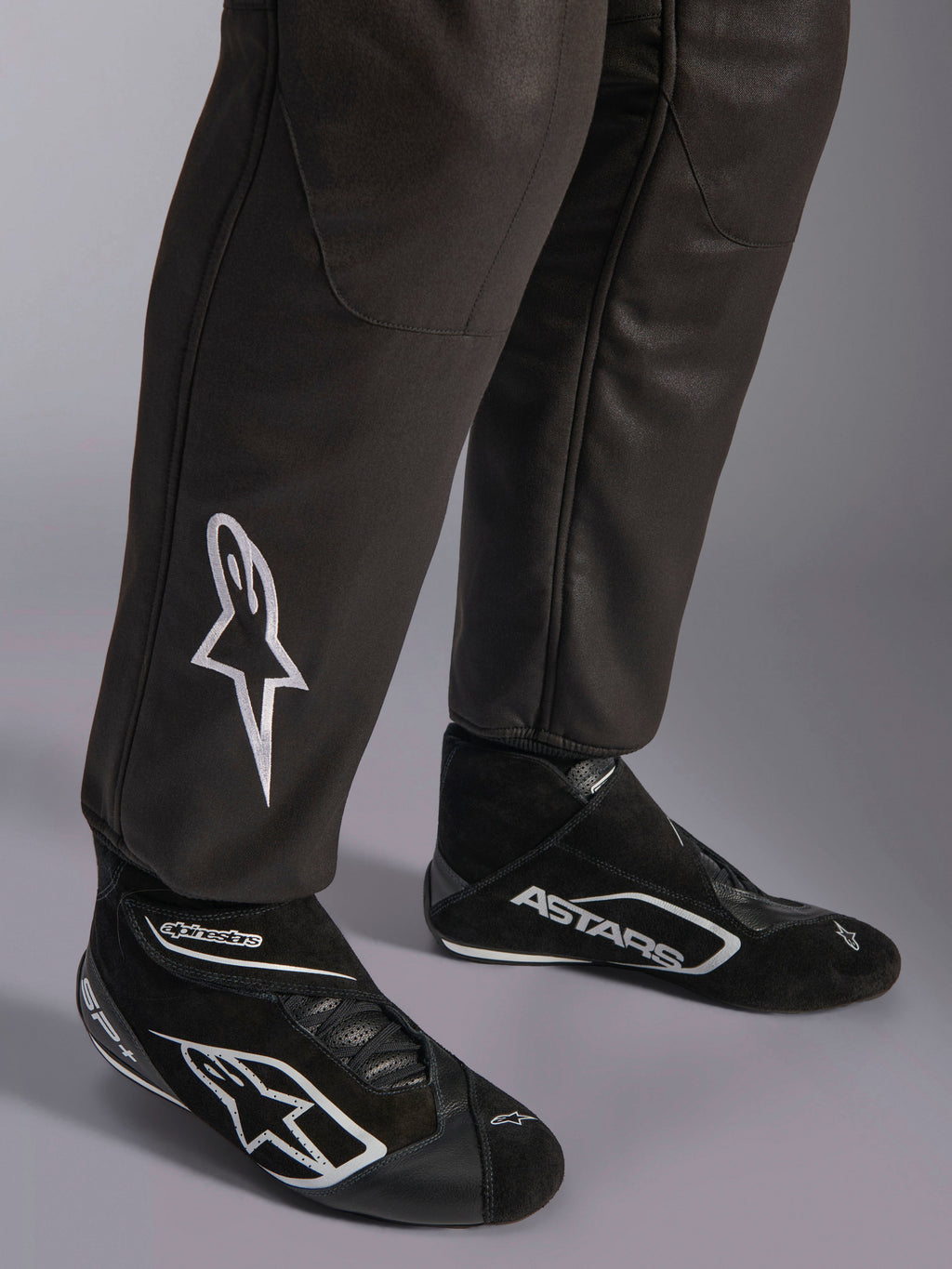 Alpinestars GP Race V3 FIA Anzug, Nahaufnahme der Unterschenkel und Rennstiefel, Schwarz mit silbernen Akzenten, weiß besticktes Stern-Logo auf dem Schienbein, gerippte Knöchelbündchen, professionelle Automobil-Rennsport-Schutzausrüstung