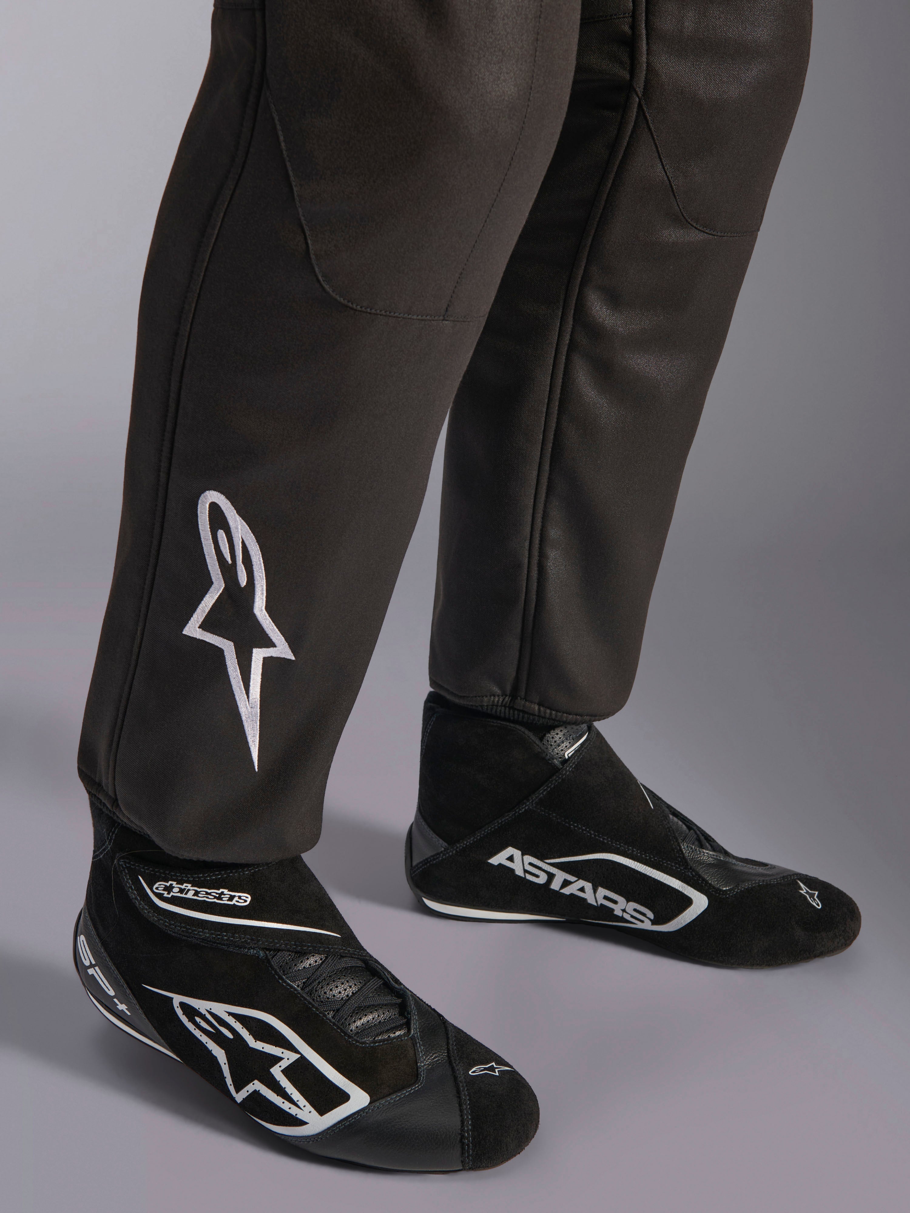 Alpinestars GP Race V3 FIA Anzug, Nahaufnahme der Unterschenkel und Rennstiefel, Schwarz mit silbernen Akzenten, weiß besticktes Stern-Logo auf dem Schienbein, gerippte Knöchelbündchen, professionelle Automobil-Rennsport-Schutzausrüstung