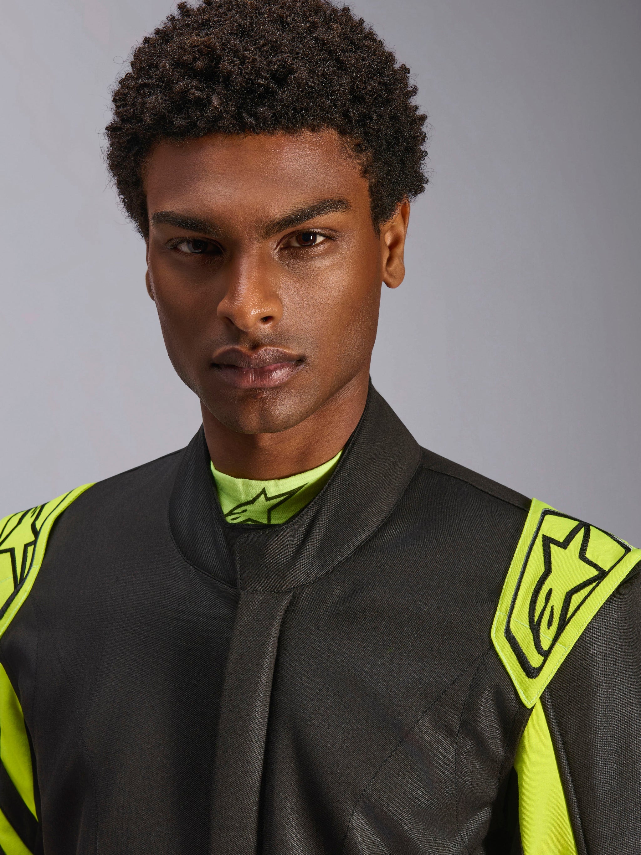 Close-up of a model wearing the Alpinestars GP Race V3 Suit FIA in Black and Yellow Fluo, featuring yellow extraction epaulettes on the shoulders with star logos, high collar design, professional auto racing gear.