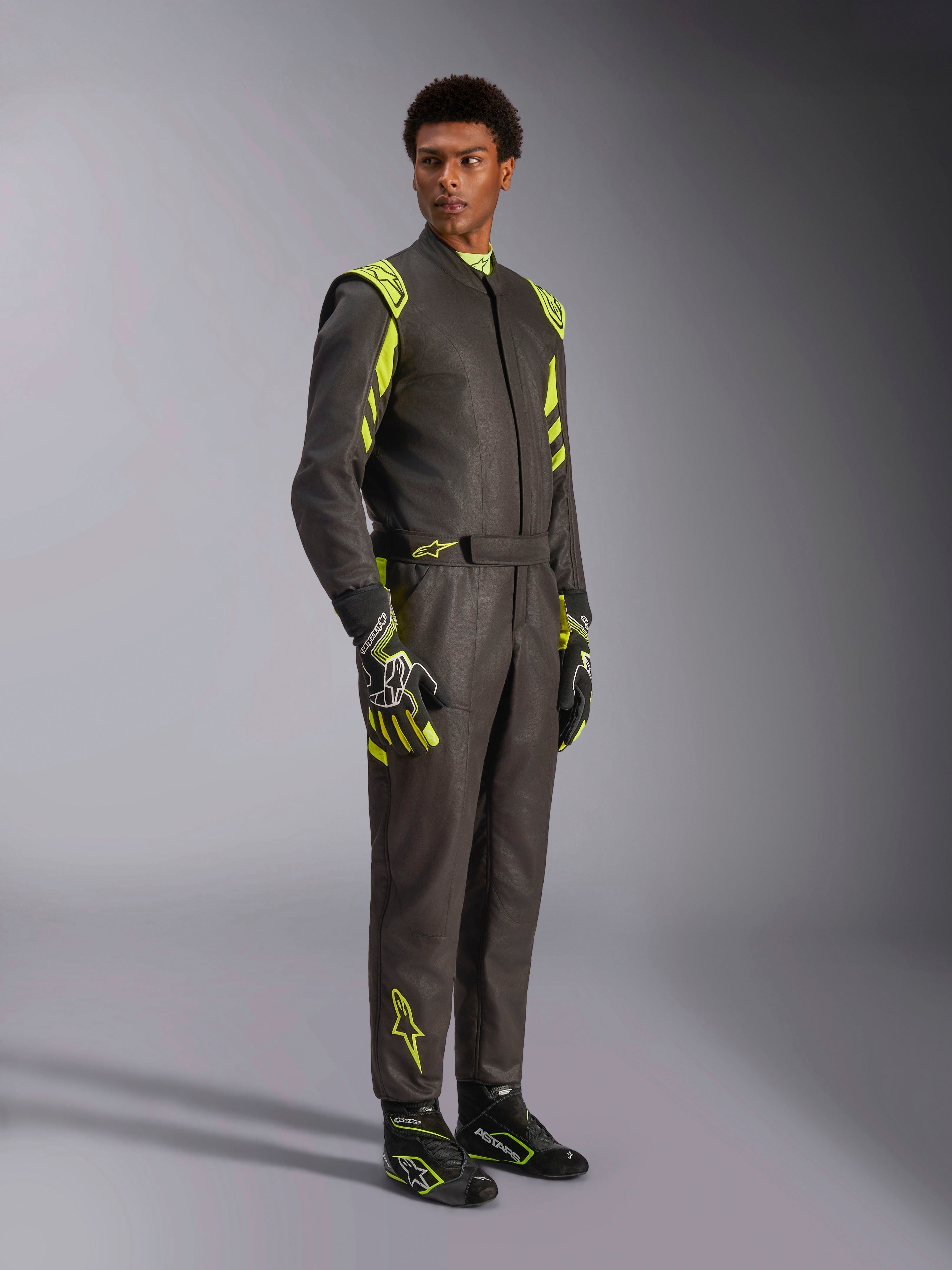 Alpinestars GP Race V3 Anzug FIA, Auto-Rennanzug, Schwarz und Fluo-Gelb, getragen von einem Model mit passenden Rennhandschuhen und Stiefeln, mit leuchtend gelben Akzenten an Schultern und Beinen, professionelle Motorsport-Ausrüstung