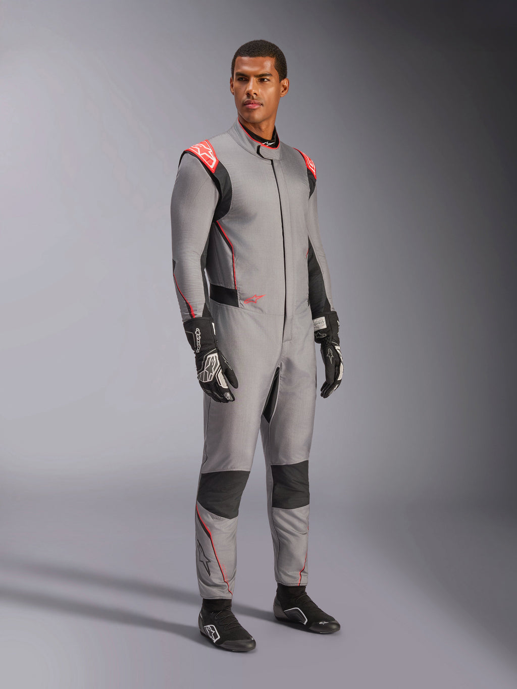 Male model wearing the Alpinestars Hypertech V3 Suit in Mid Gray with Black and Red Fluo accents, professional auto racing suit paired with black racing gloves and shoes, featuring a lightweight two-layer Nomex construction with red shoulder detailing and an athletic fit