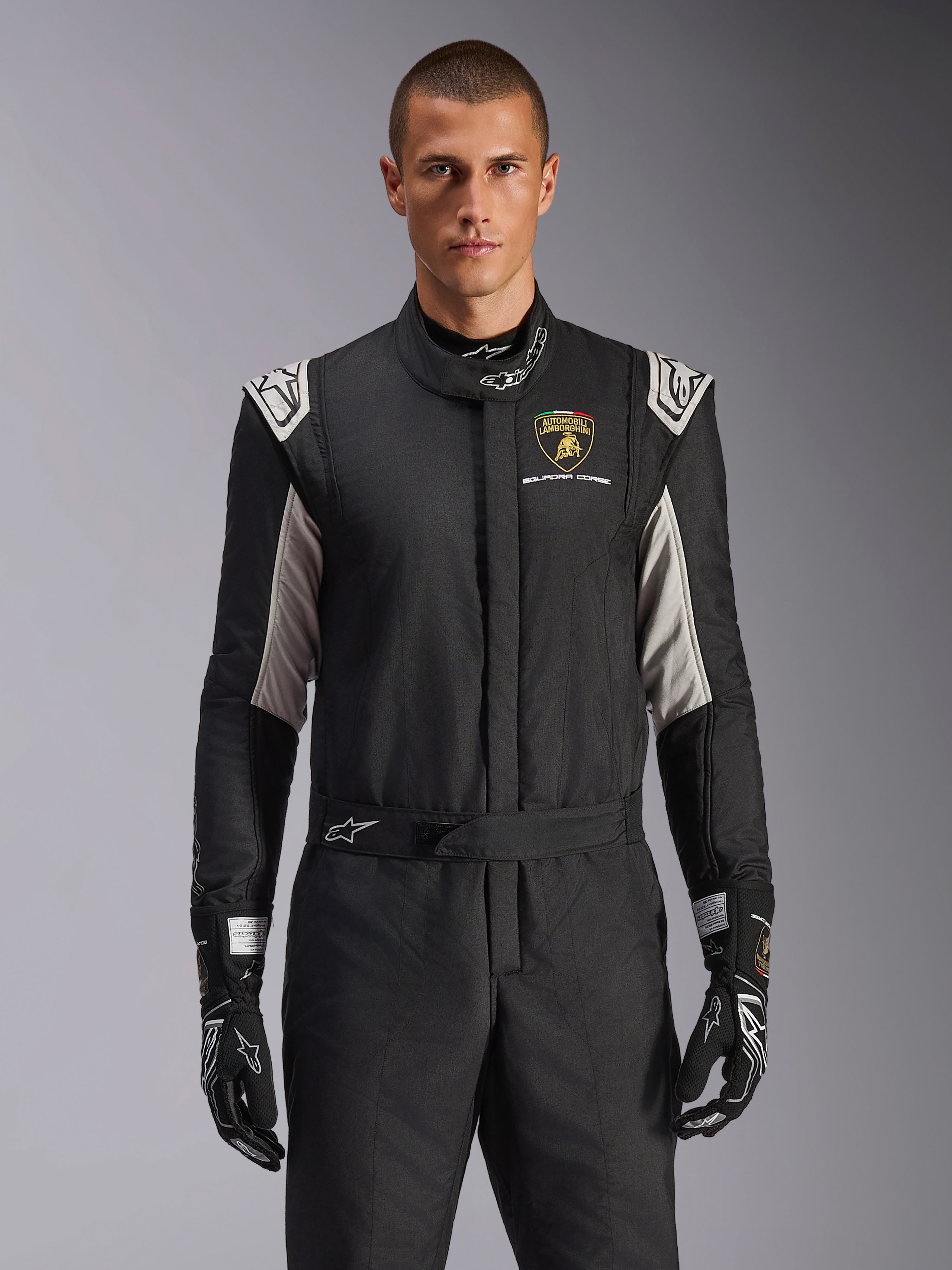 Alpinestars Limited Edition Lamborghini GP Tech V4 Suit, professional racing suit in black with silver and gold accents, featuring the Automobili Lamborghini Squadra Corse shield on the chest, worn by a model with matching black racing gloves.