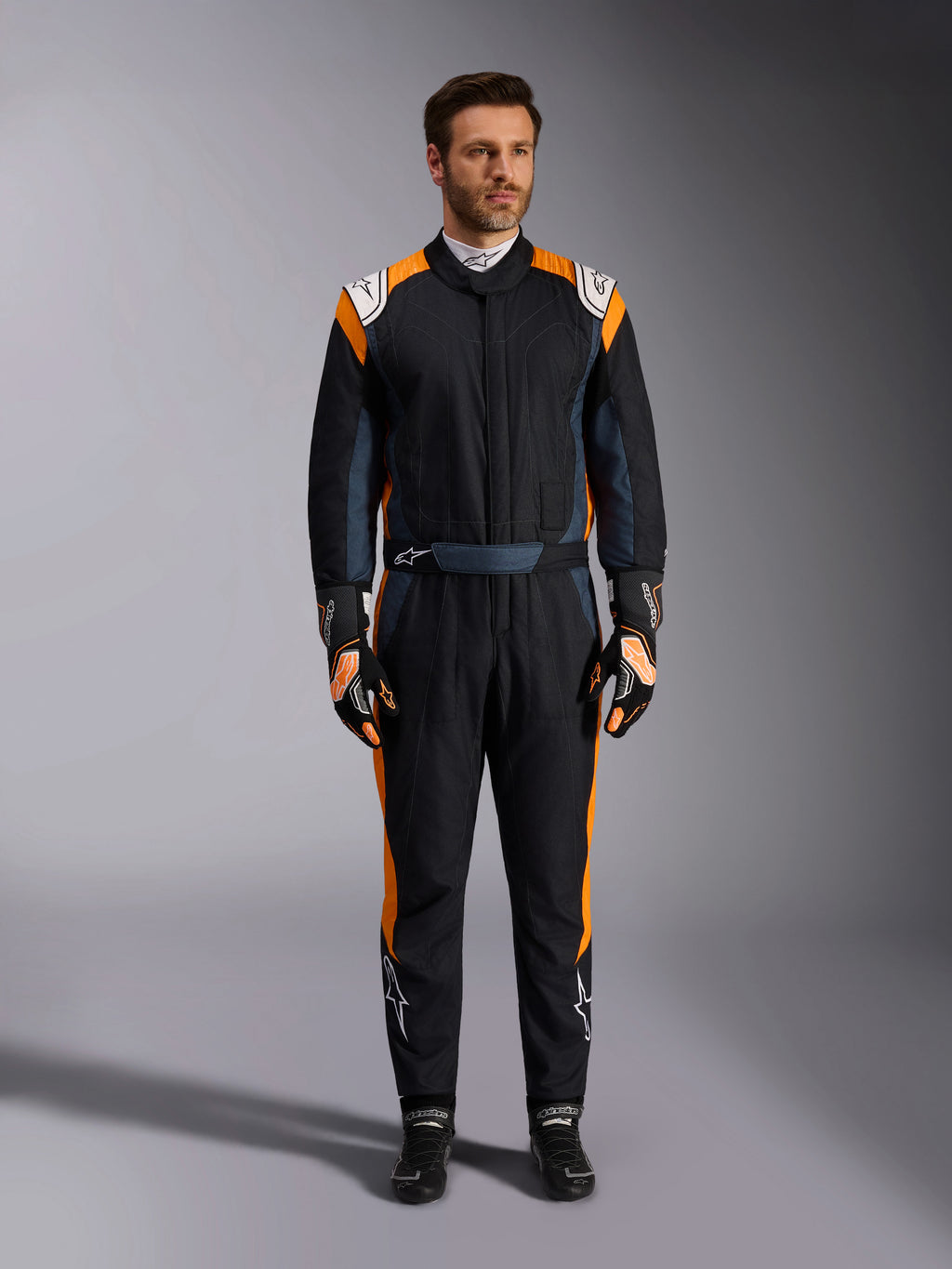 Alpinestars GP Pro Comp V2 FIA Rennanzug, Autorennanzug in Schwarz Asphalt und Orange Fluo, männliches Model in vollständiger Schutzausrüstung mit passenden Rennhandschuhen und Schuhen, weiße Alpinestars-Logos und leuchtend orangefarbene Seitenpaneele, professionelle Motorsport-Bekleidung