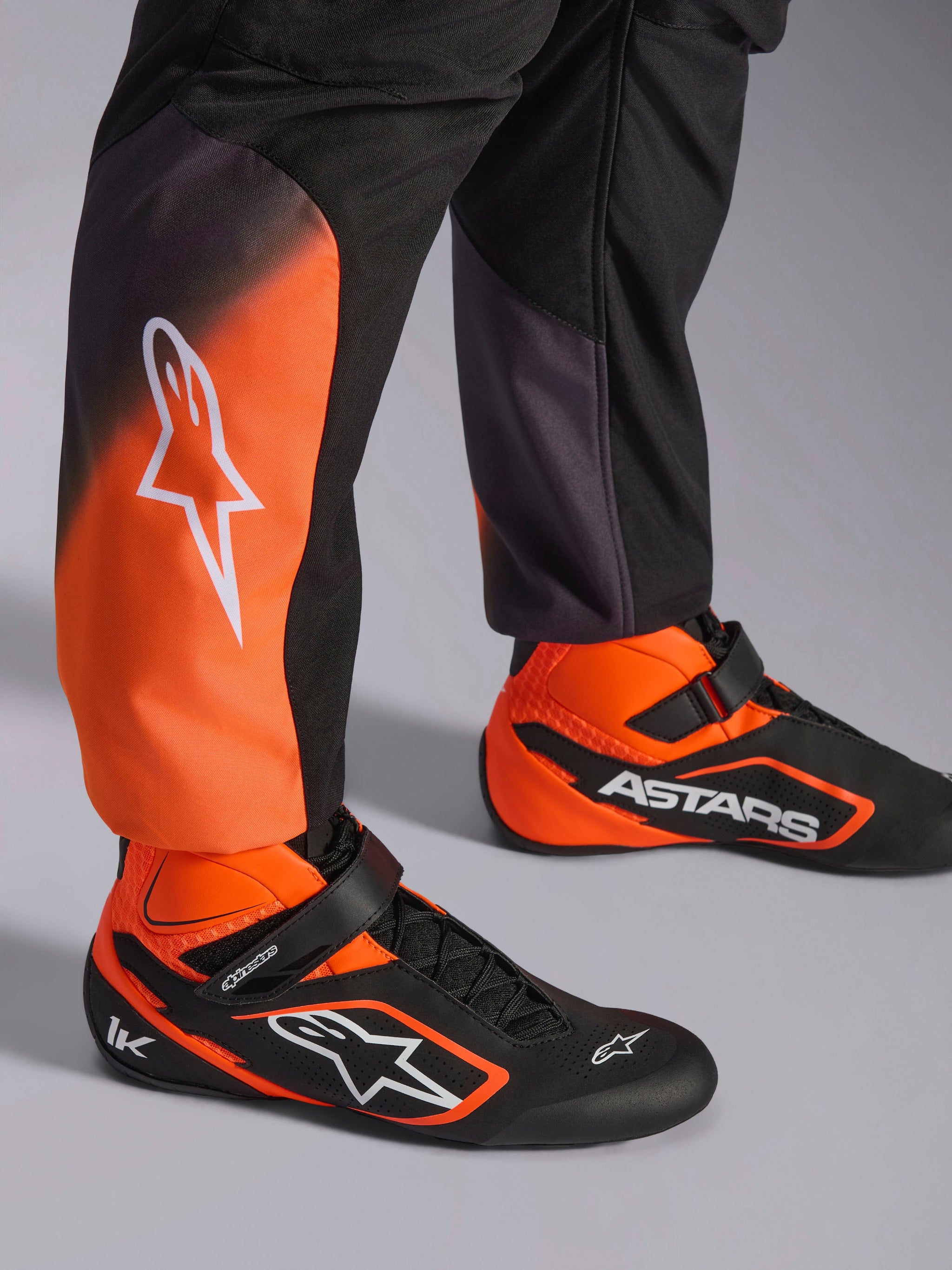 Close-up of Alpinestars KMX-5 V3 Suit legs, Black and Orange Fluo karting suit, featuring white logo on the calf, paired with matching black and orange professional racing shoes