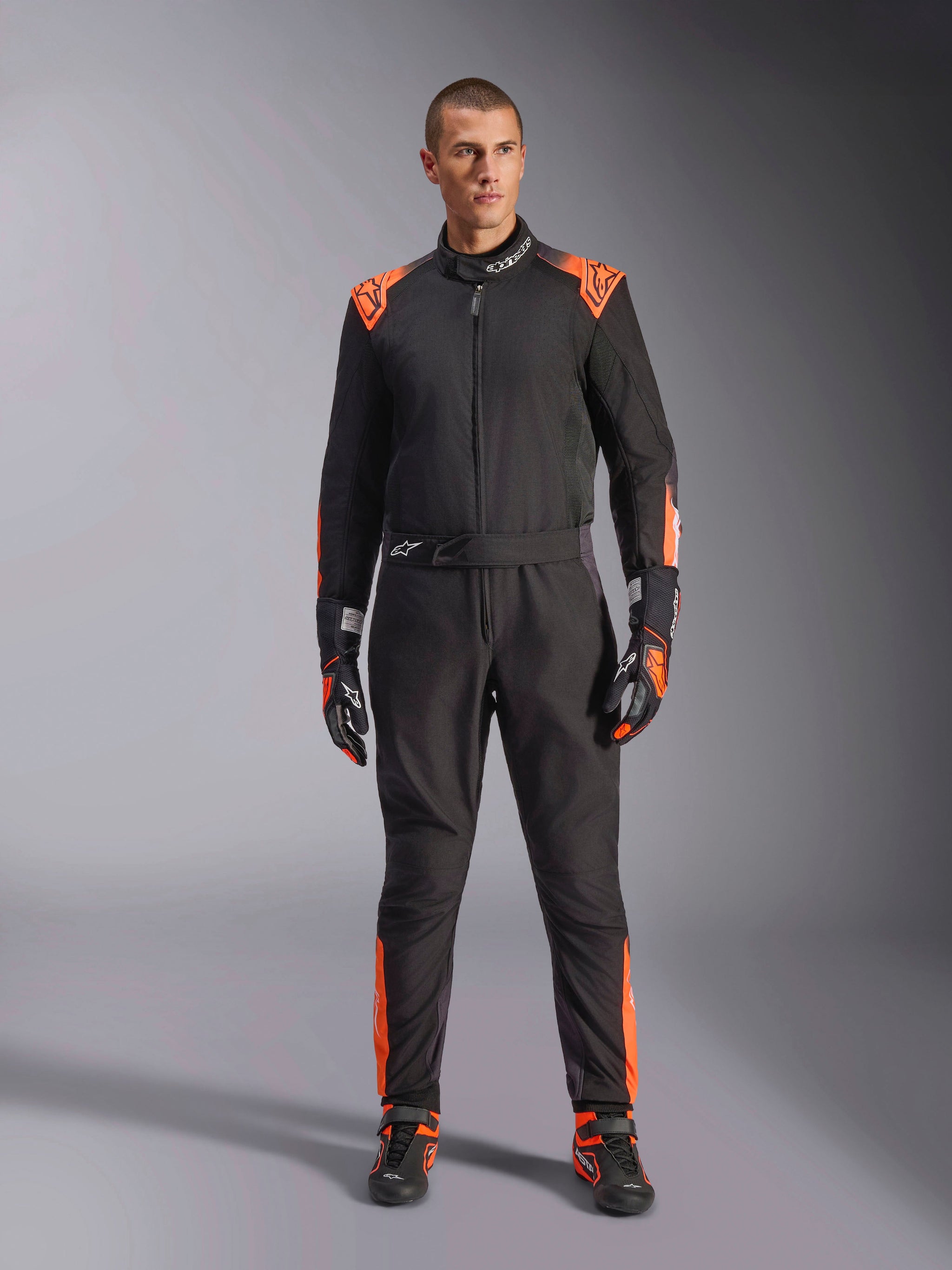Alpinestars KMX-5 V3 Suit, Black and Orange Fluo karting suit, worn by a model with matching racing gloves and shoes, featuring orange accents on the shoulders and legs, professional one-piece motorsport design with a technical mesh appearance.