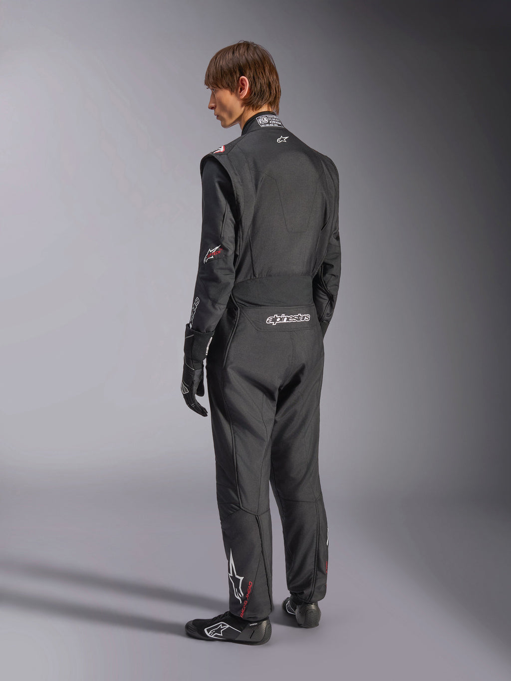 Alpinestars GP Tech V4 Suit, Auto Racing Suit, Black with white accents, back view of model wearing the suit with racing gloves and shoes, featuring white branding on the lower back and leg, professional motorsport safety gear