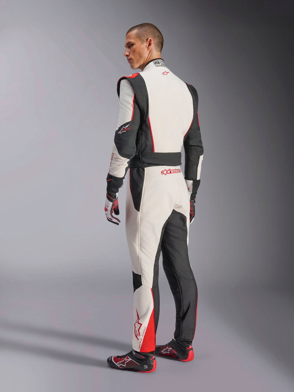 Alpinestars GP Tech V4 Suit, White Black Red racing suit, rear view showing ergonomic stretch panels on the lower back and legs, paired with matching racing gloves and shoes, professional motorsport apparel.