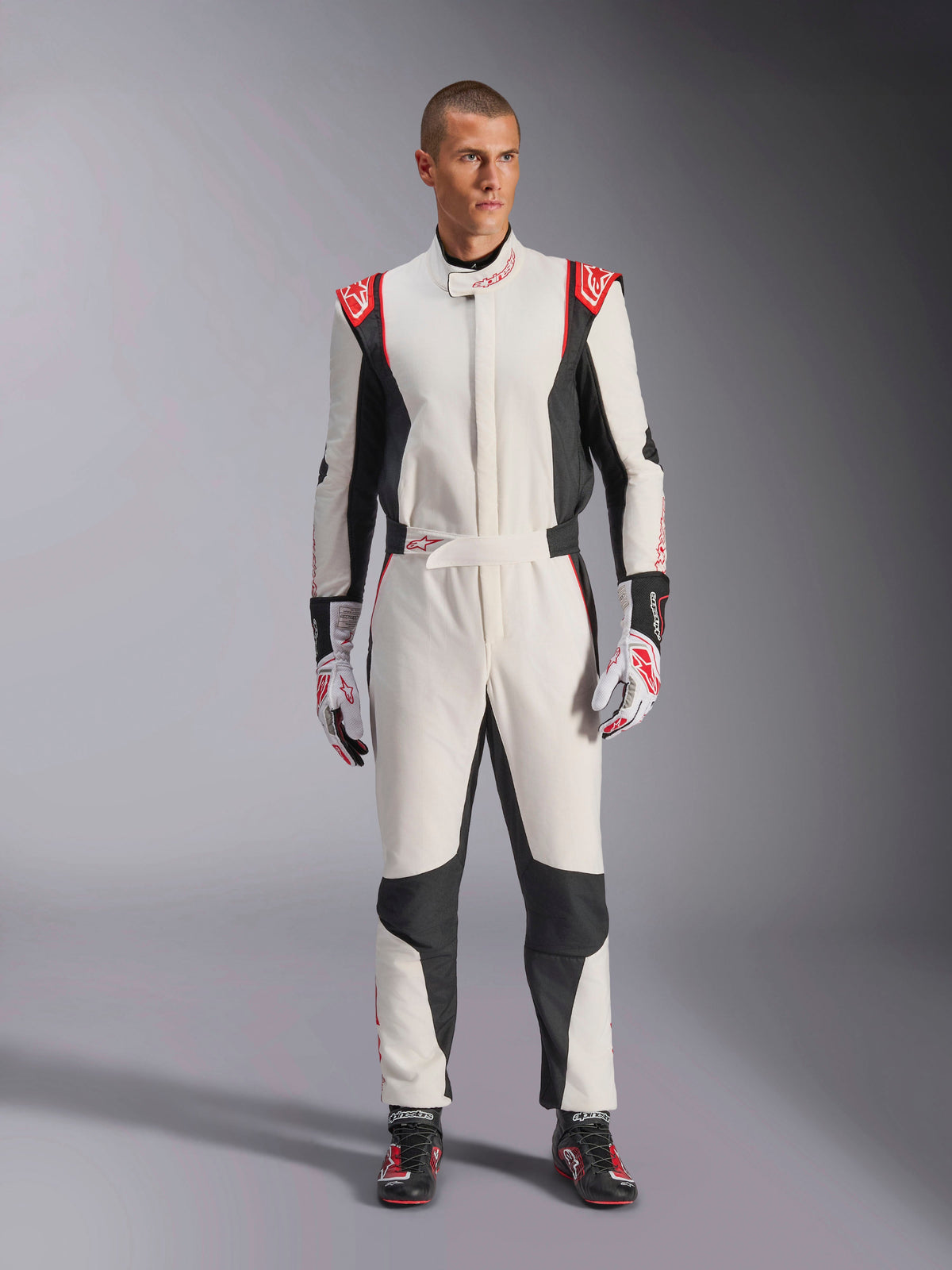 Alpinestars GP Tech V4 Anzug, einteiliger Autorennanzug in Weiß, Schwarz und Rot, getragen von einem männlichen Model mit passenden Rennhandschuhen und -schuhen, mit roten Logos auf den Schultern und schwarzen Seiteneinsätzen, professionelle Motorsport-Ausrüstung für die Rennstrecke.