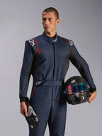Male model wearing Alpinestars GP Tech V4 Suit, Navy Blue auto racing suit with black and red accents, holding a carbon fiber helmet and racing gloves, professional motorsport protective gear