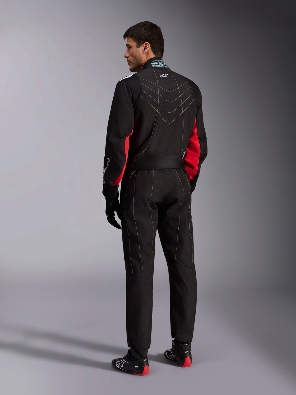 Alpinestars KMX-9 V3 Anzug, Schwarz Rot Weiß, einteiliger Kart-Anzug in der Rückansicht, mit weißen geometrischen Nähten am Rücken und roten Akzenten an den Ärmeln, getragen mit schwarzen Rennhandschuhen und Kart-Schuhen.