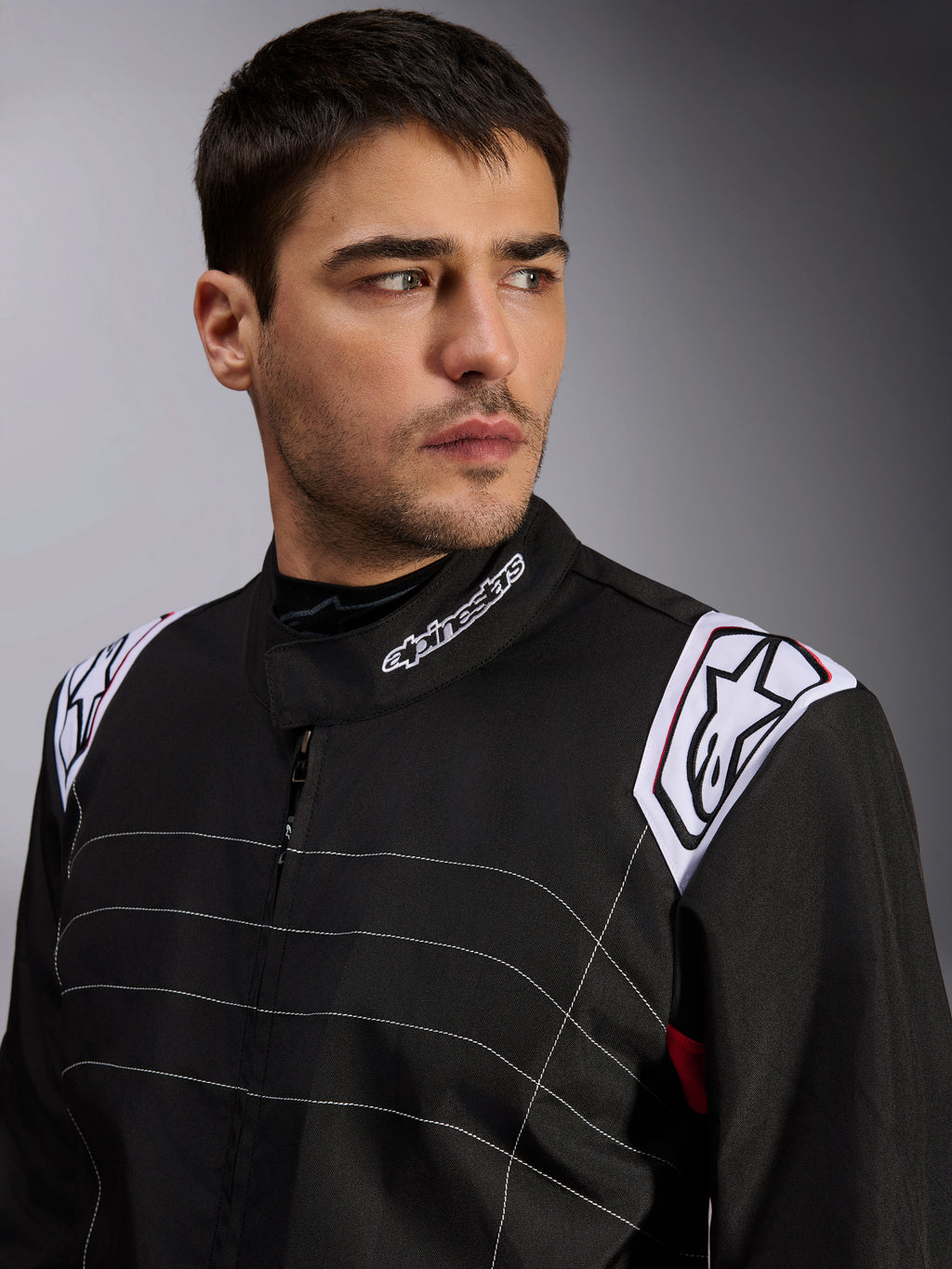 Alpinestars KMX-9 V3 Anzug, schwarzer Kart-Anzug, getragen von einem männlichen Model, Nahaufnahme von Brust und Schultern mit weißem Branding am Kragen und an den Epauletten, mit weißen Ziernähten und einem professionellen Motorsport-Design